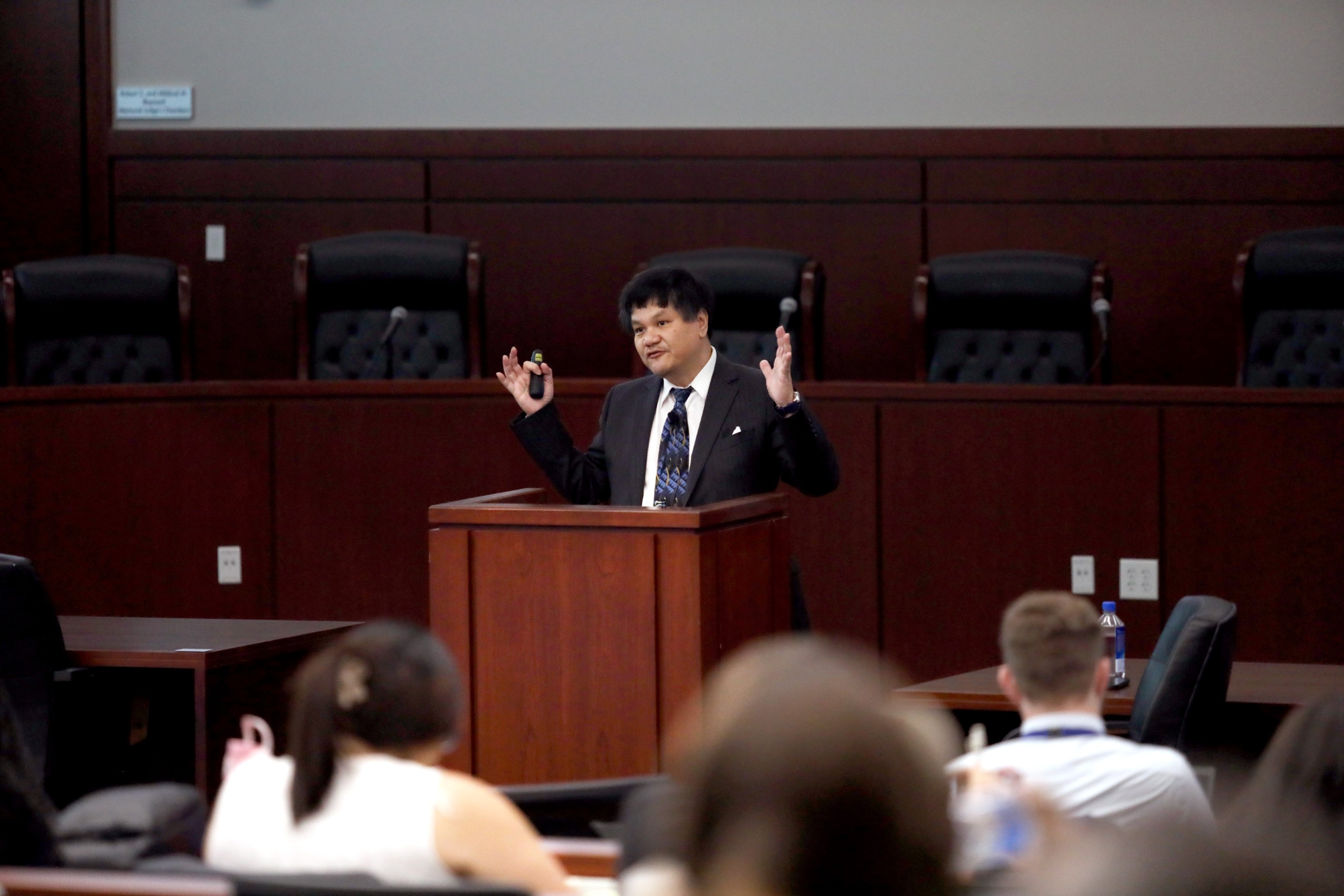 A person is speaking behind a lectern in a large room filled with seated audience members, some of whom are holding their phones or taking notes.