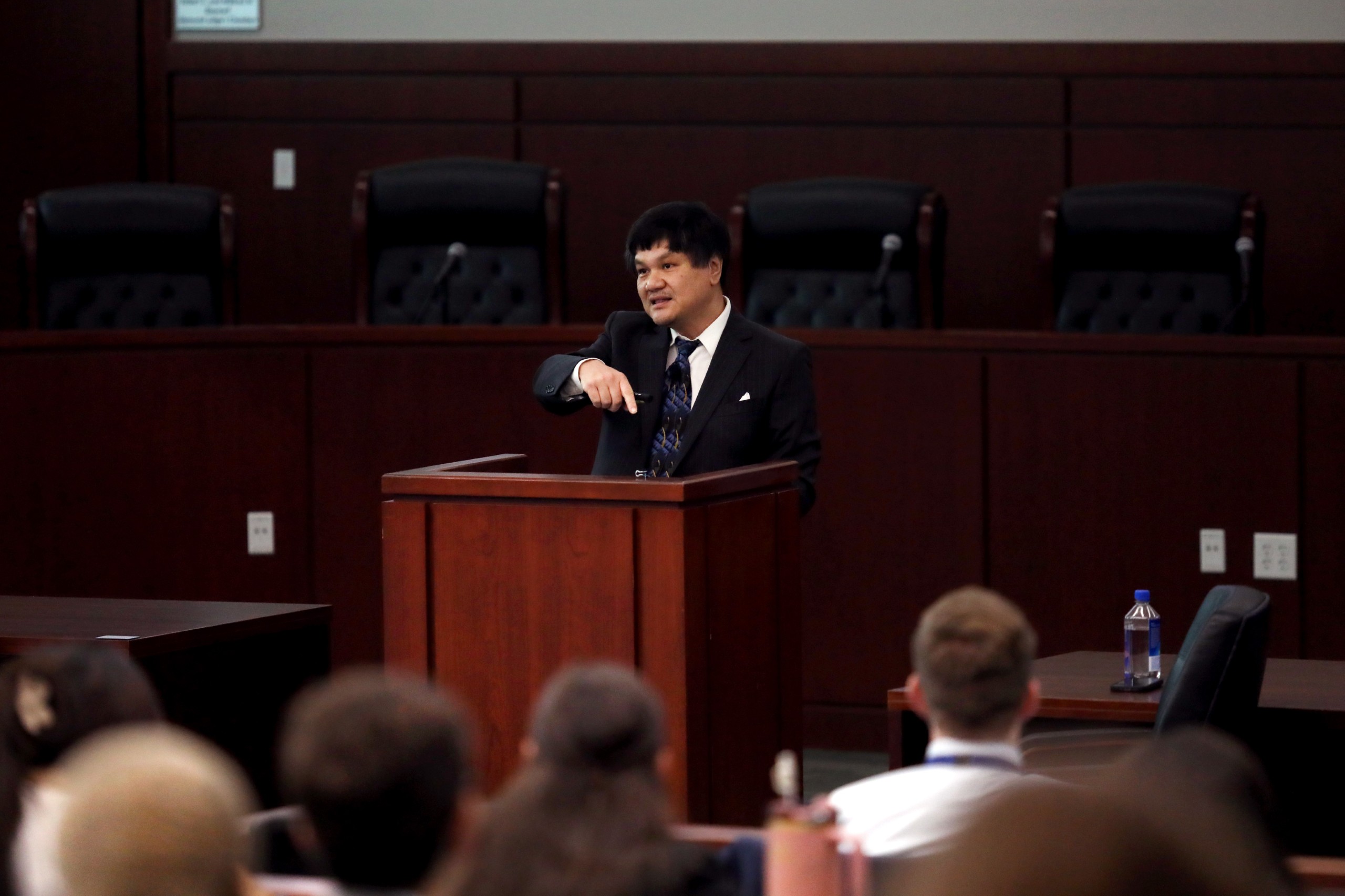 A person stands behind a lectern speaking to an audience in a conference setting. The audience is seated, facing the speaker.