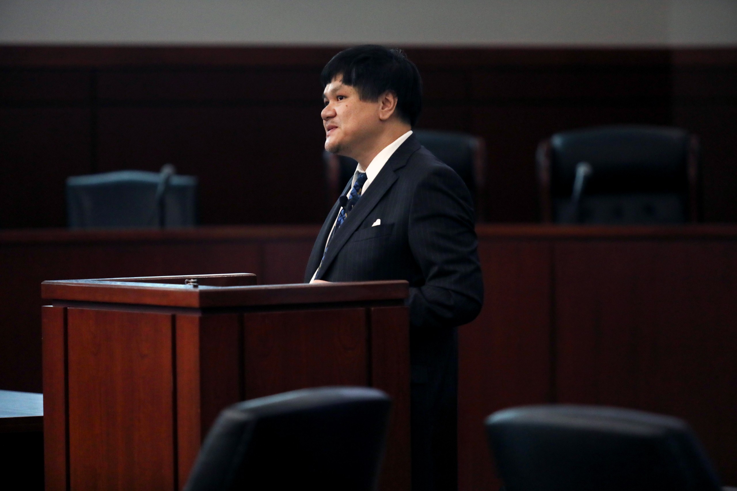 A person speaks behind a lectern in a formal setting, with empty chairs visible in the background.