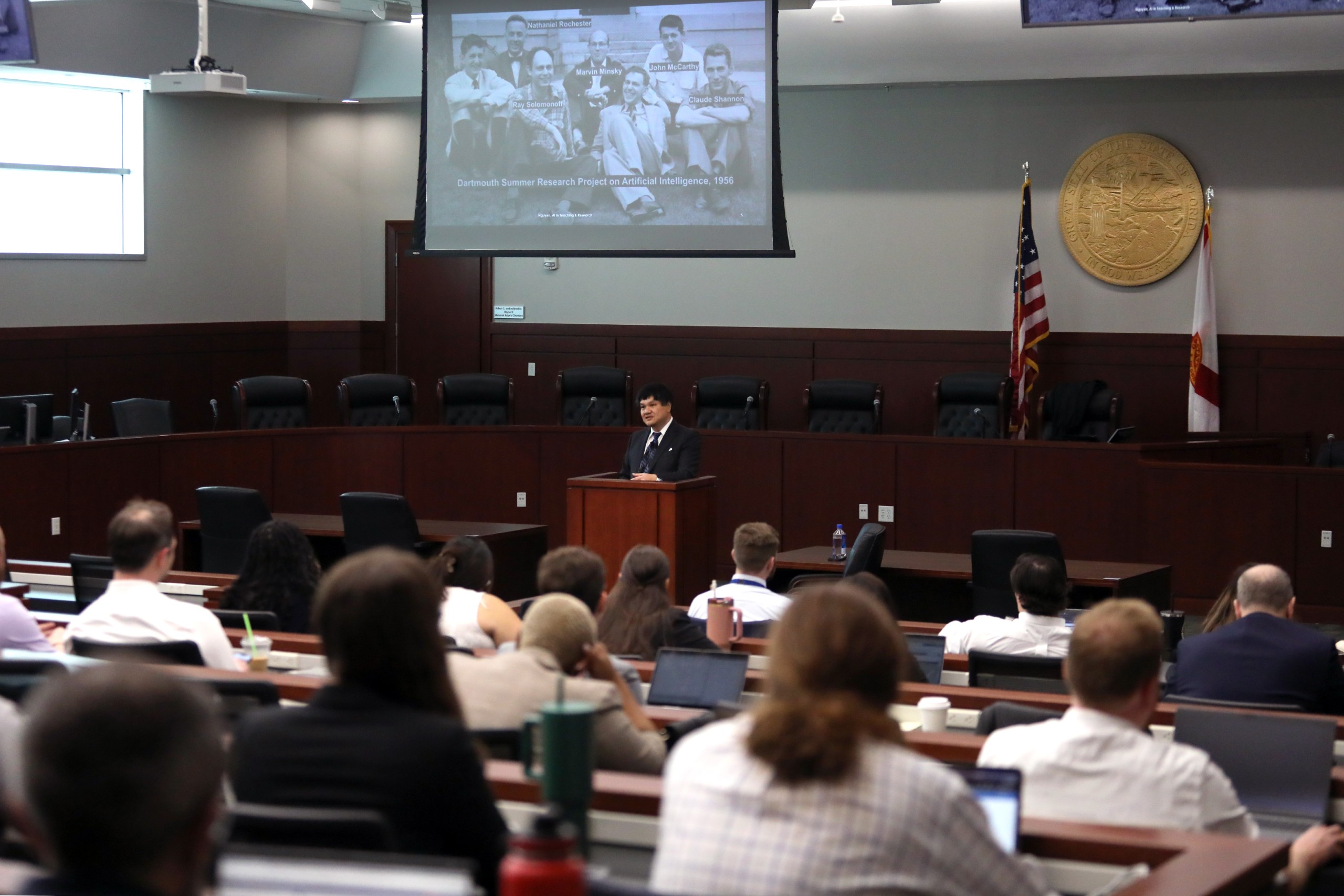 A person stands behind a lectern speaking to an audience in a meeting room. In the background, a screen displays a historical photo related to artificial intelligence. Several attendees are seated, some with laptops and beverages.
