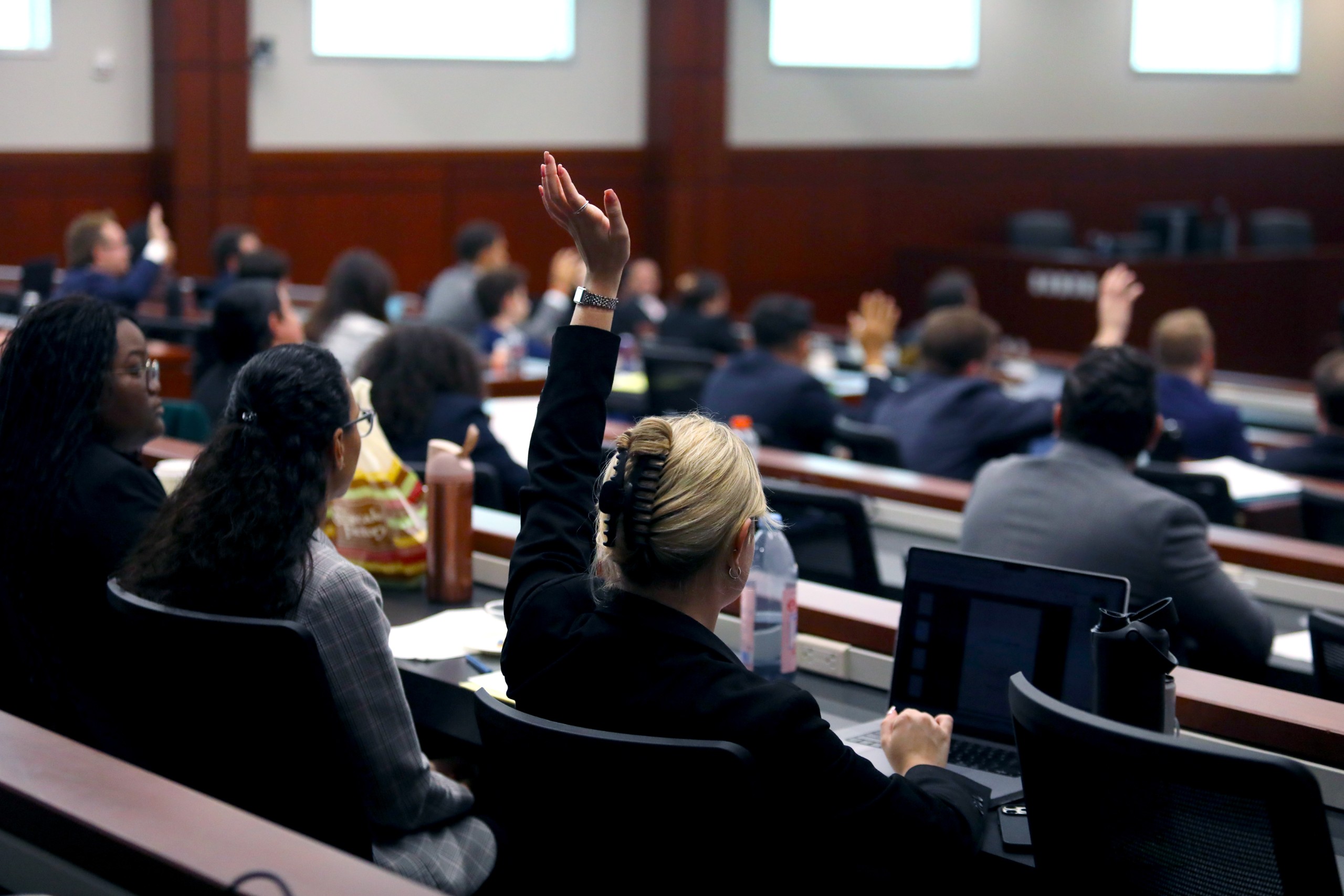 An individual seated in a courtroom, dressed in legal attire, appears relaxed while holding an object in their hand. The background includes desks and equipment typically found in a legal setting.