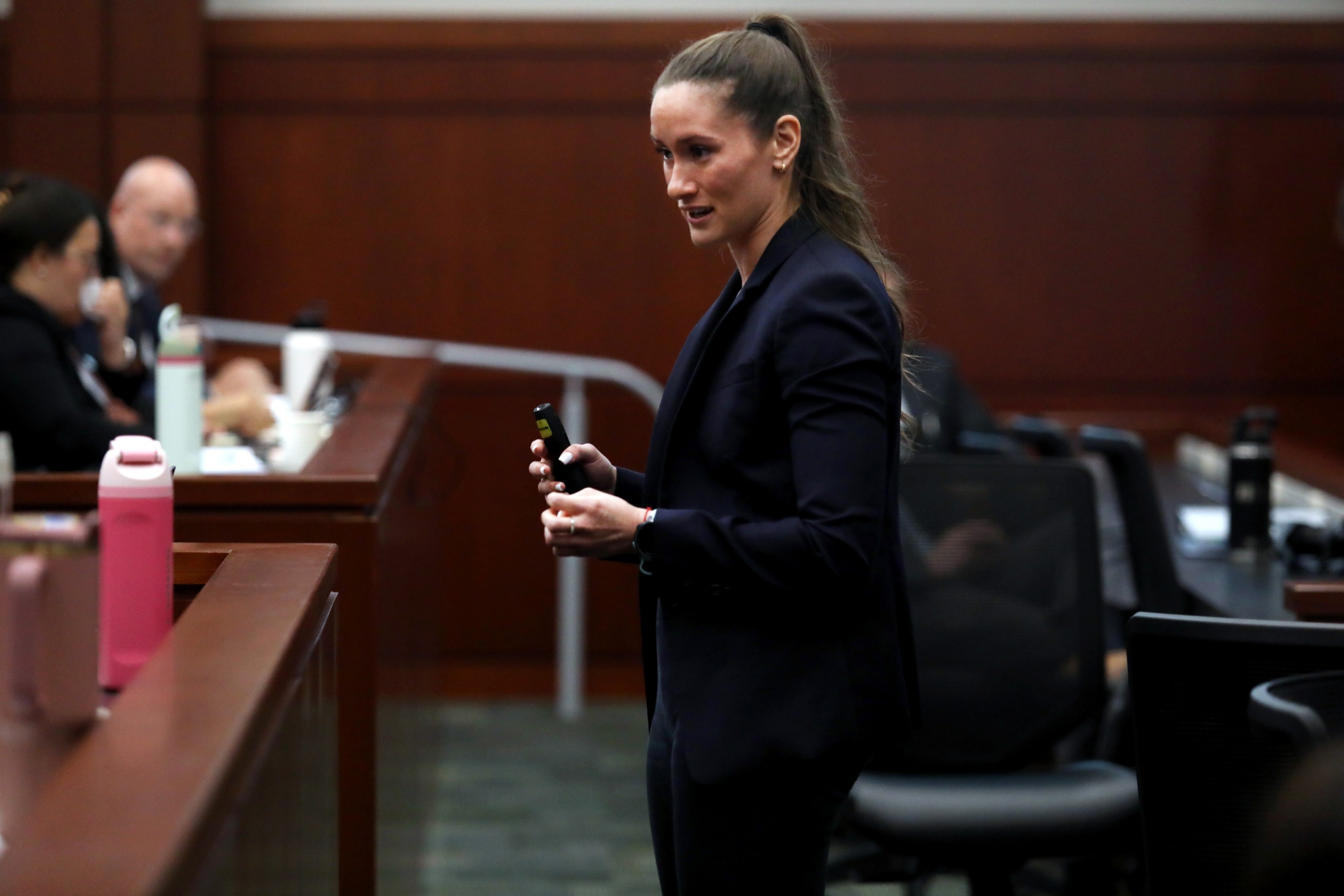 A person stands behind a lectern in a courtroom, holding a small device and addressing an audience. Several individuals are seated in the background, facing the speaker.