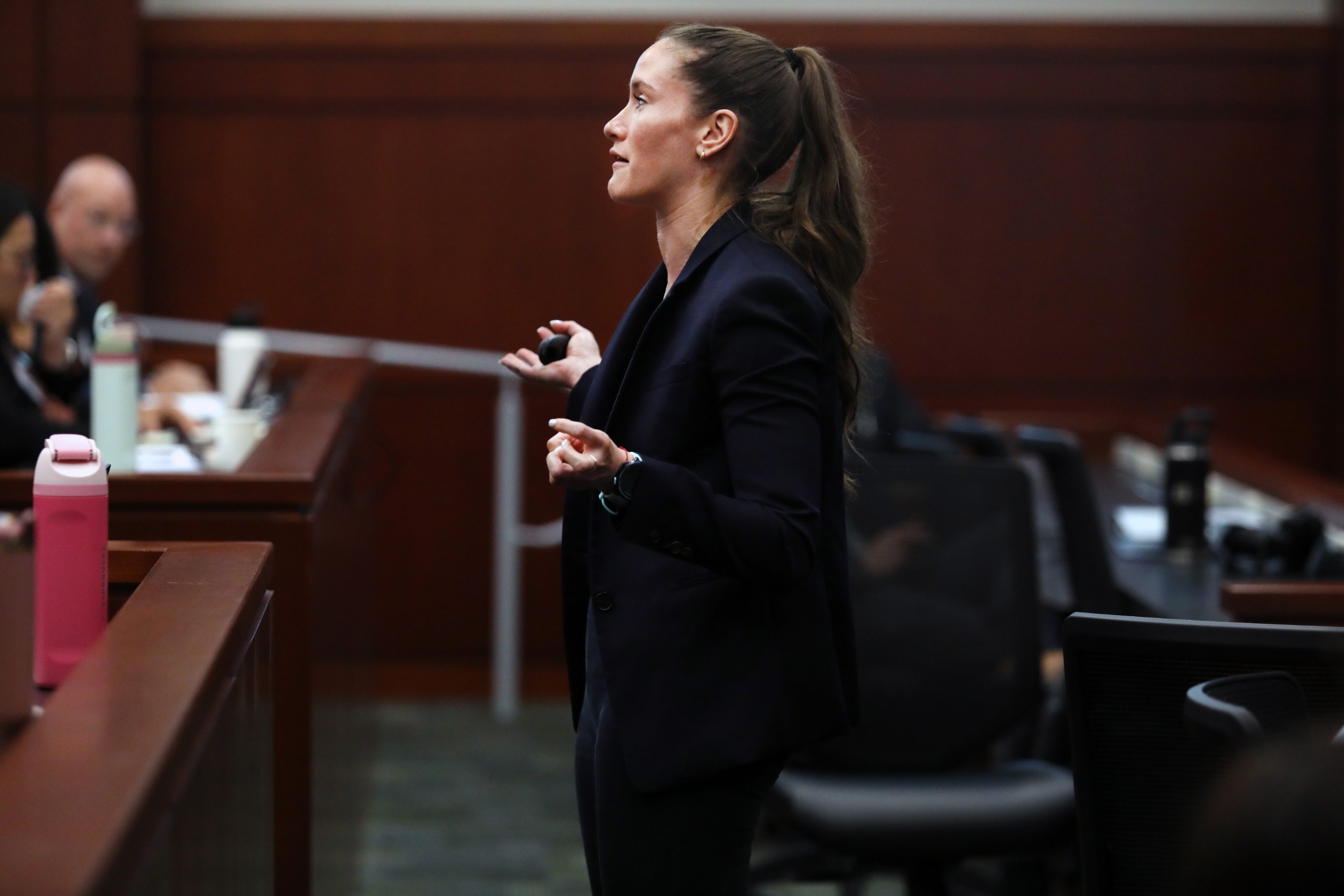 An individual stands speaking into a microphone, addressing an audience. They are positioned in a formal setting with wood-paneled walls, while a few seated attendees listen attentively.