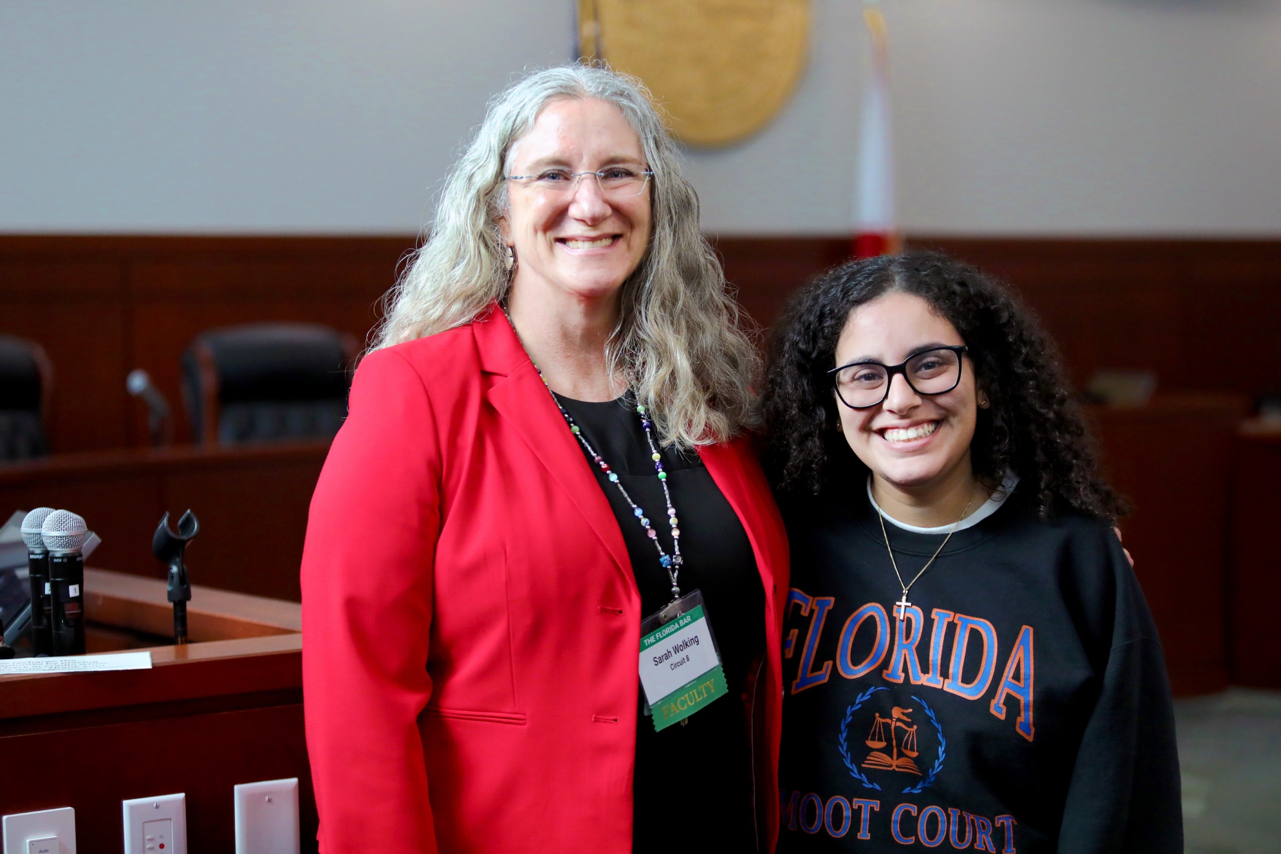 Two individuals are standing together, smiling for the camera. One is wearing a red blazer and a name tag, while the other is in a sweatshirt that says "Florida Moot Court." They are positioned in a courtroom setting, with chairs and a flag visible in the background.