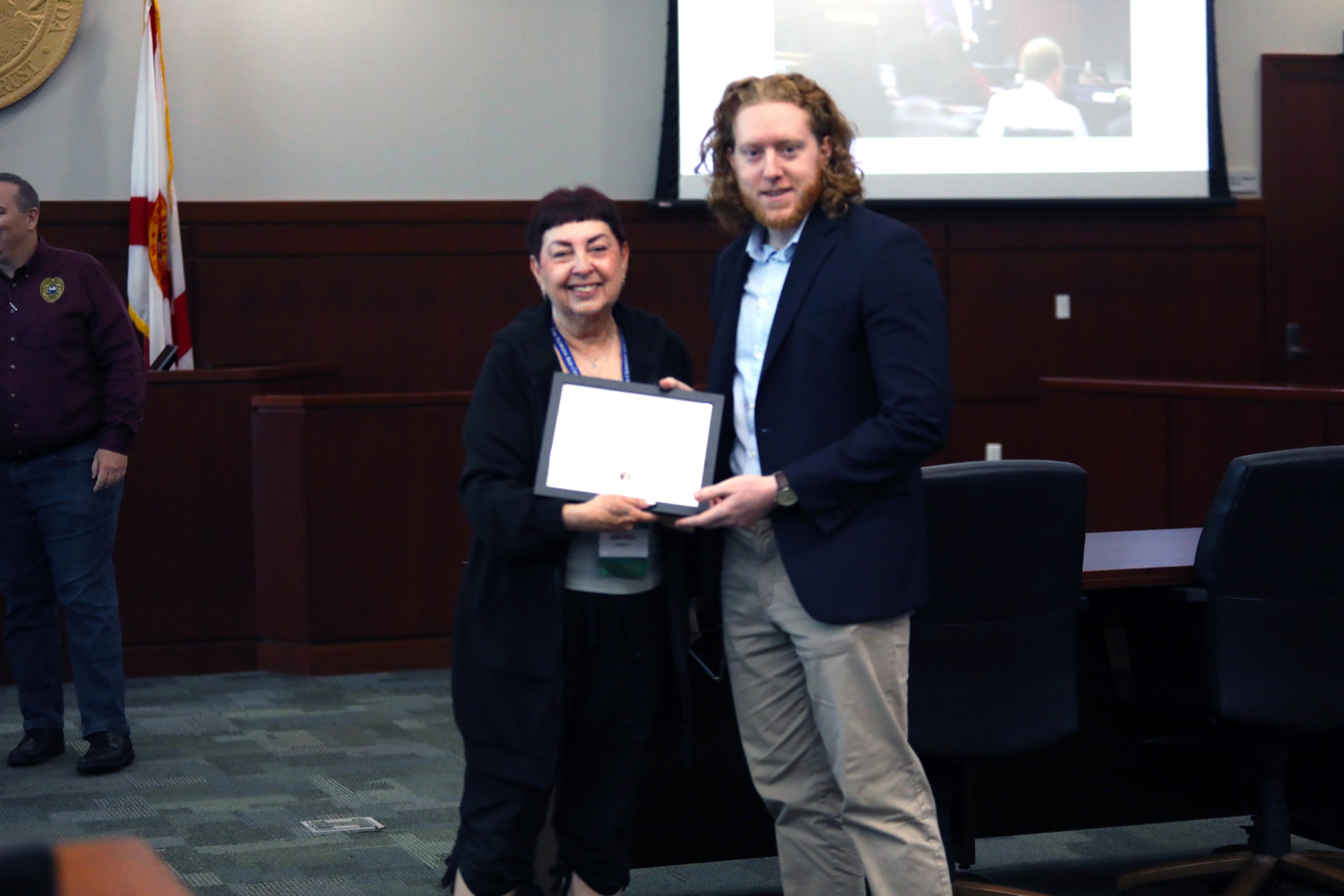 Two individuals are smiling and holding a certificate together in a formal setting. One person is in the foreground, while another is seen in the background, standing beside a flag. A screen displaying an image is visible in the background.