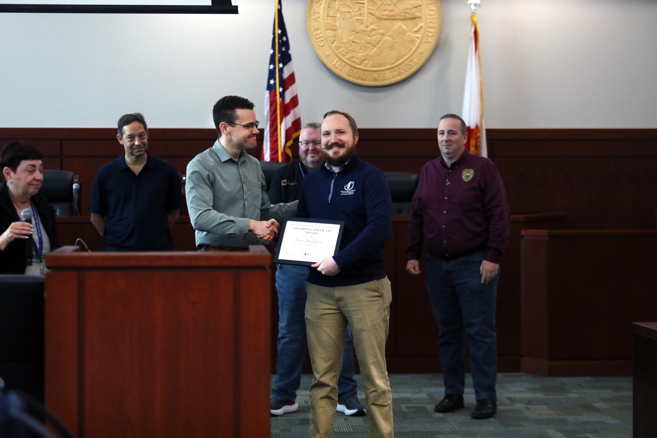 A person shakes hands with another while holding a certificate. In the background, several individuals observe the exchange. A seal and flags are visible on the wall behind them.