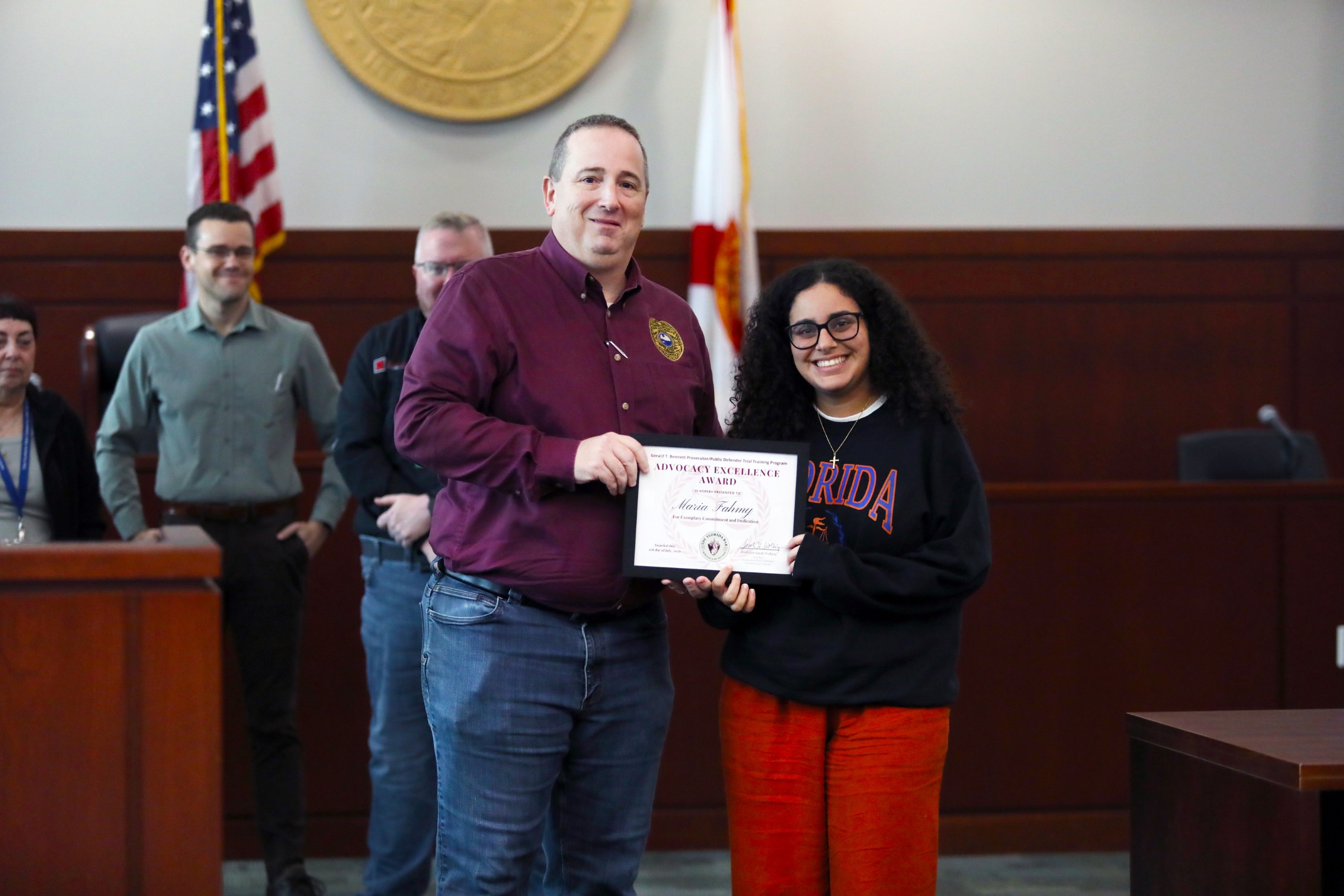 A person standing behind a lectern presents a certificate to another individual in a courtroom setting. Several people are visible in the background, observing the exchange.