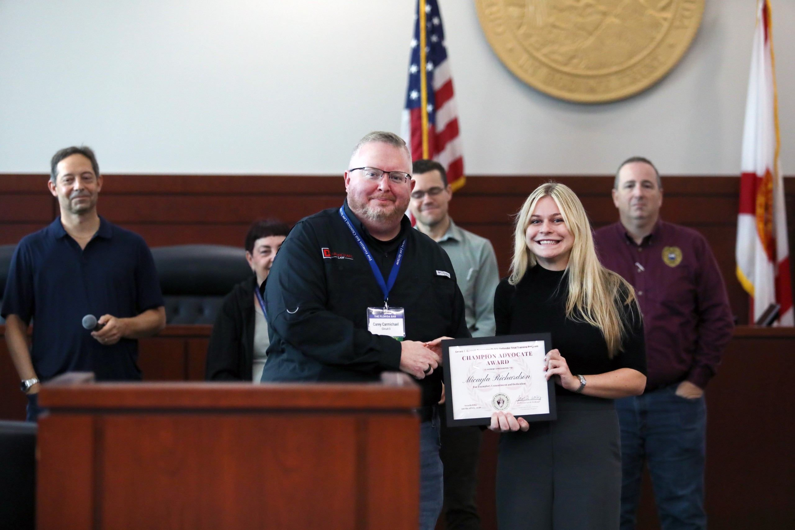 A group of individuals stands in a courtroom setting. One person is handing a certificate labeled "Champion Advocate Award" to another individual, who is smiling. Other attendees look on in the background. An American flag and a state flag are visible on either side of the scene.