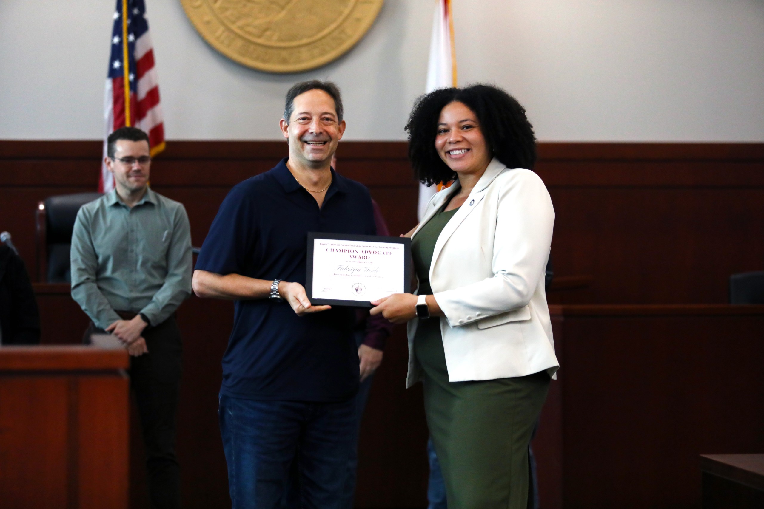 A person hands a certificate to another individual in a courtroom setting, with an audience in the background. An American flag and state flag are visible behind them.