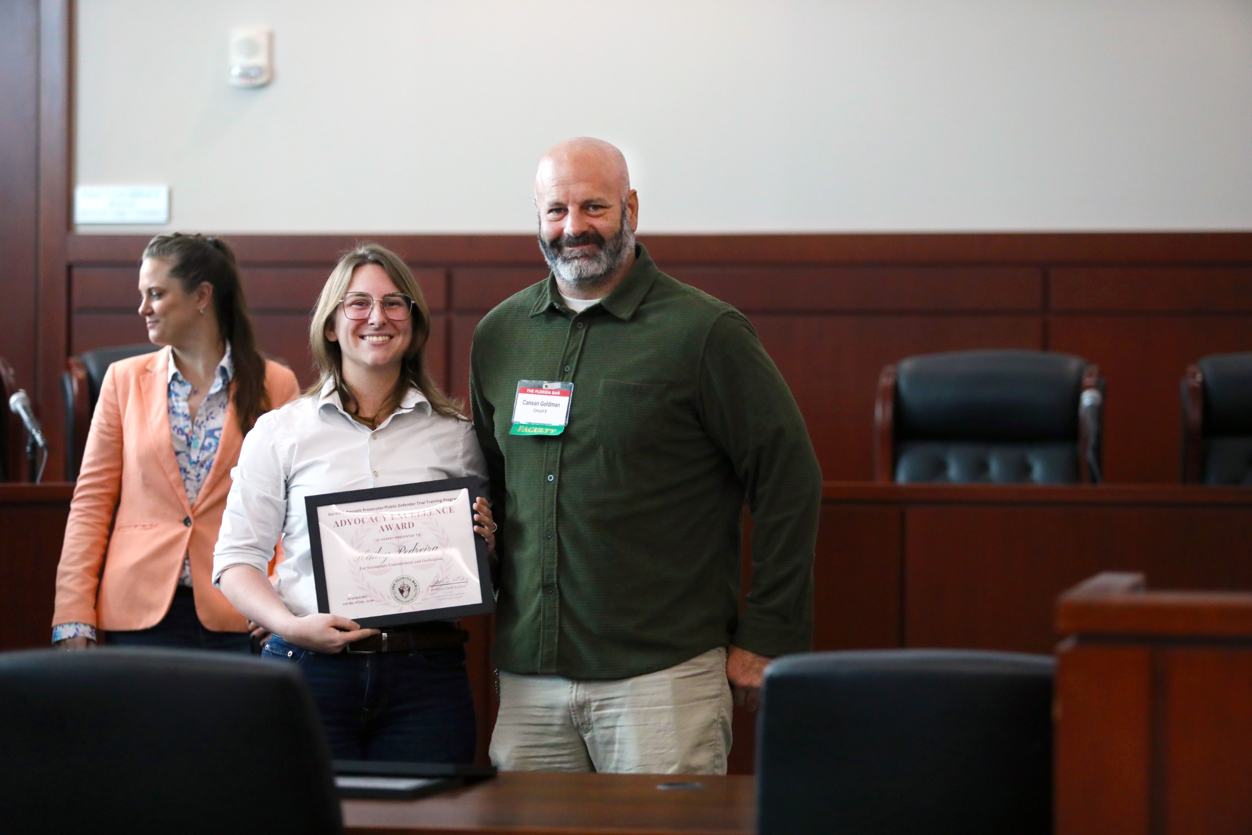 Two individuals stand in a courtroom setting, one holding a certificate. The person on the left is partially turned away, while the person on the right smiles. In the background, an empty row of chairs is visible.