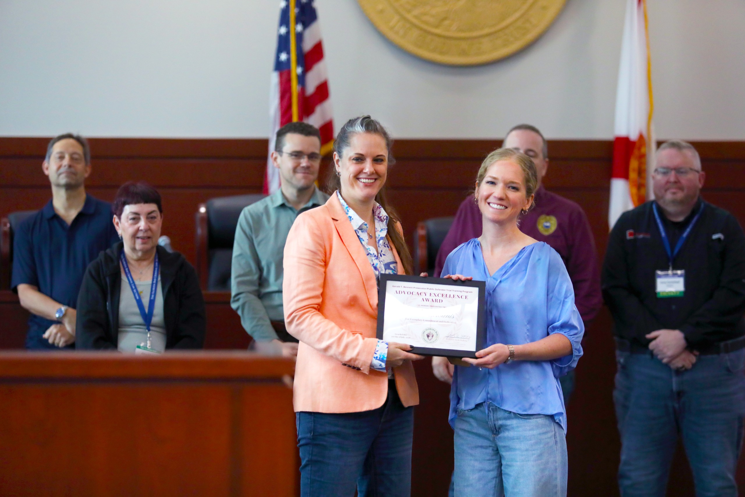 Two individuals are presenting an award certificate while standing in front of a group of people. The setting appears to be a formal event, with flags in the backdrop.