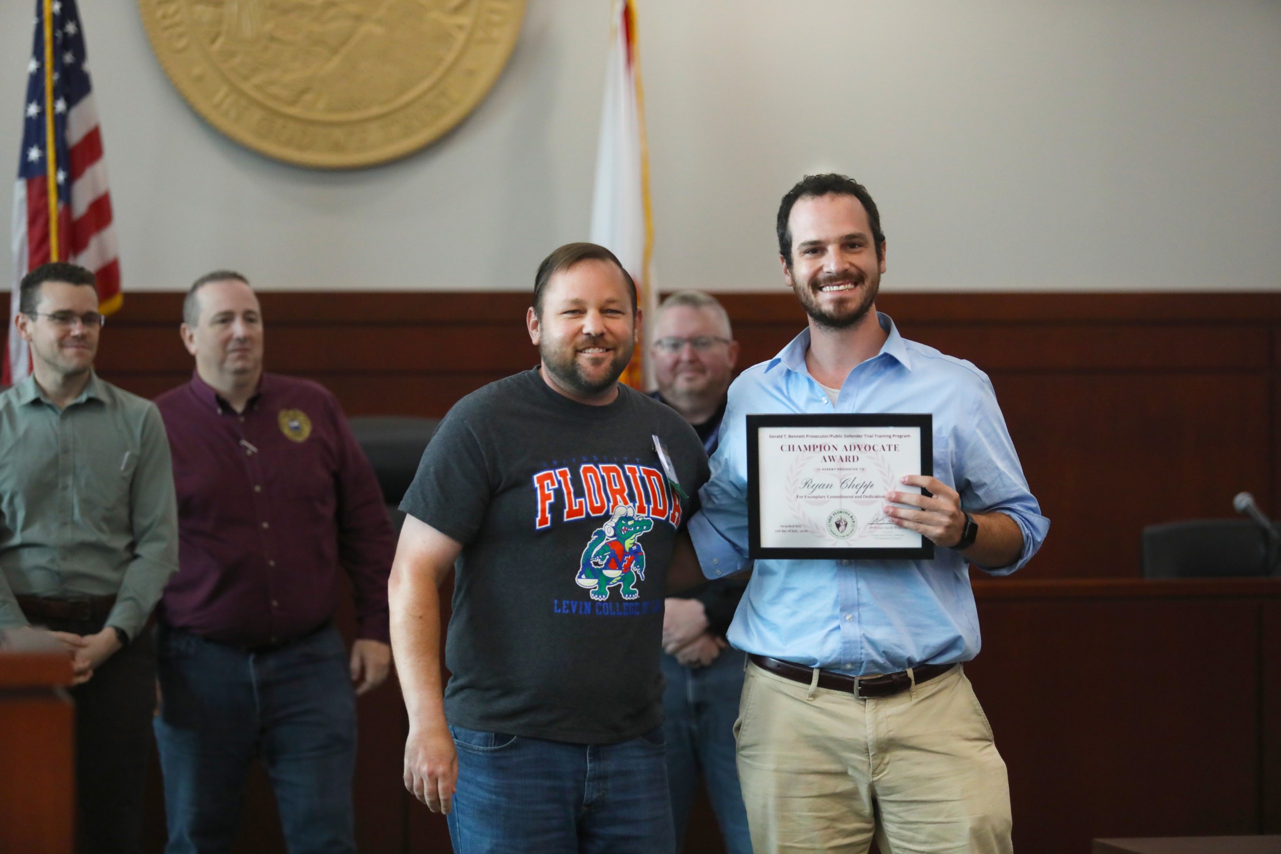 Two individuals stand in a courtroom setting. One individual holds an award certificate, while the other person, who is smiling, stands beside them. In the background, a small group of people is gathered, with flags displayed on the wall.