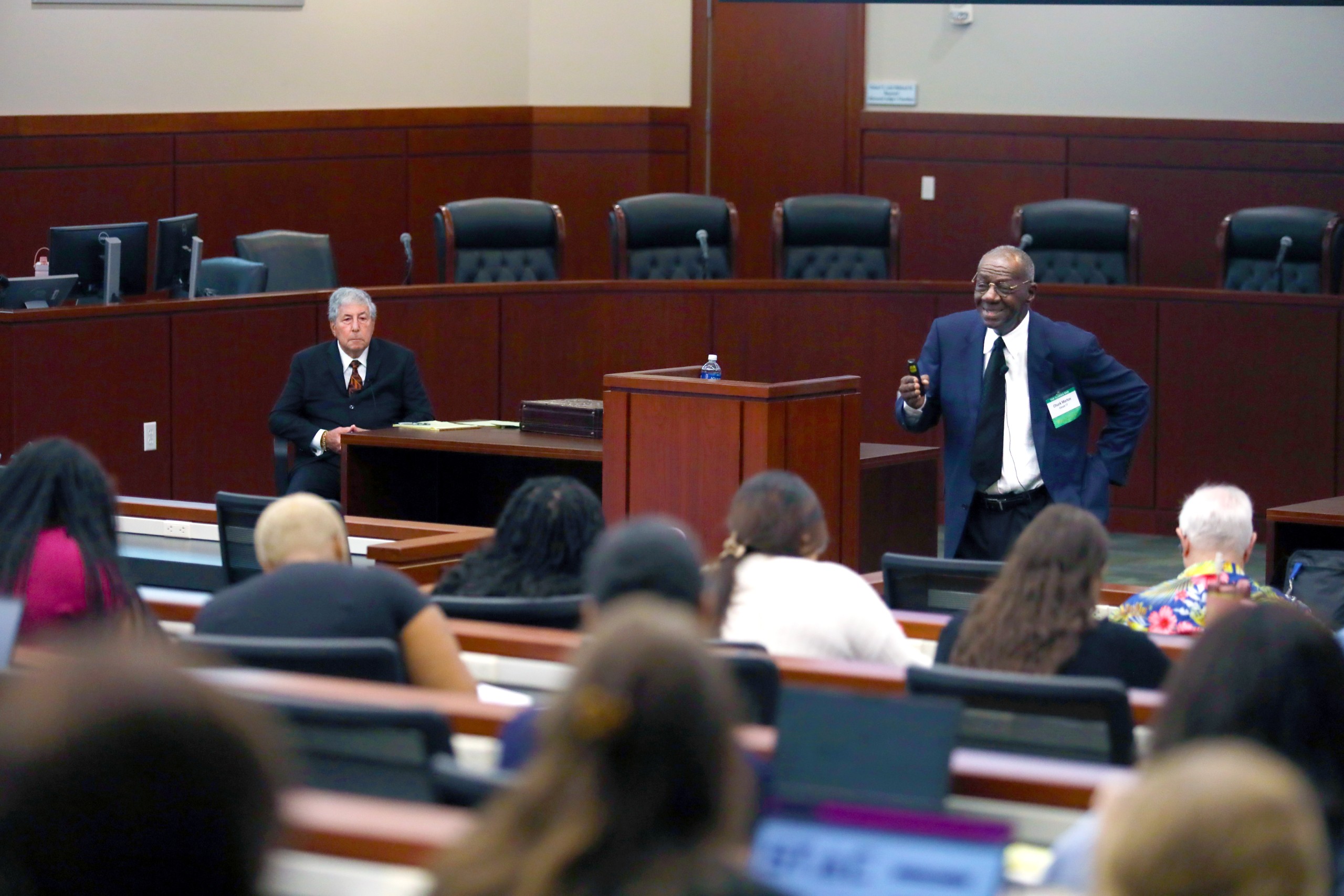 A person stands behind a lectern while speaking to an audience in a meeting room. Another individual is seated nearby, and attendees are engaged, some using laptops.