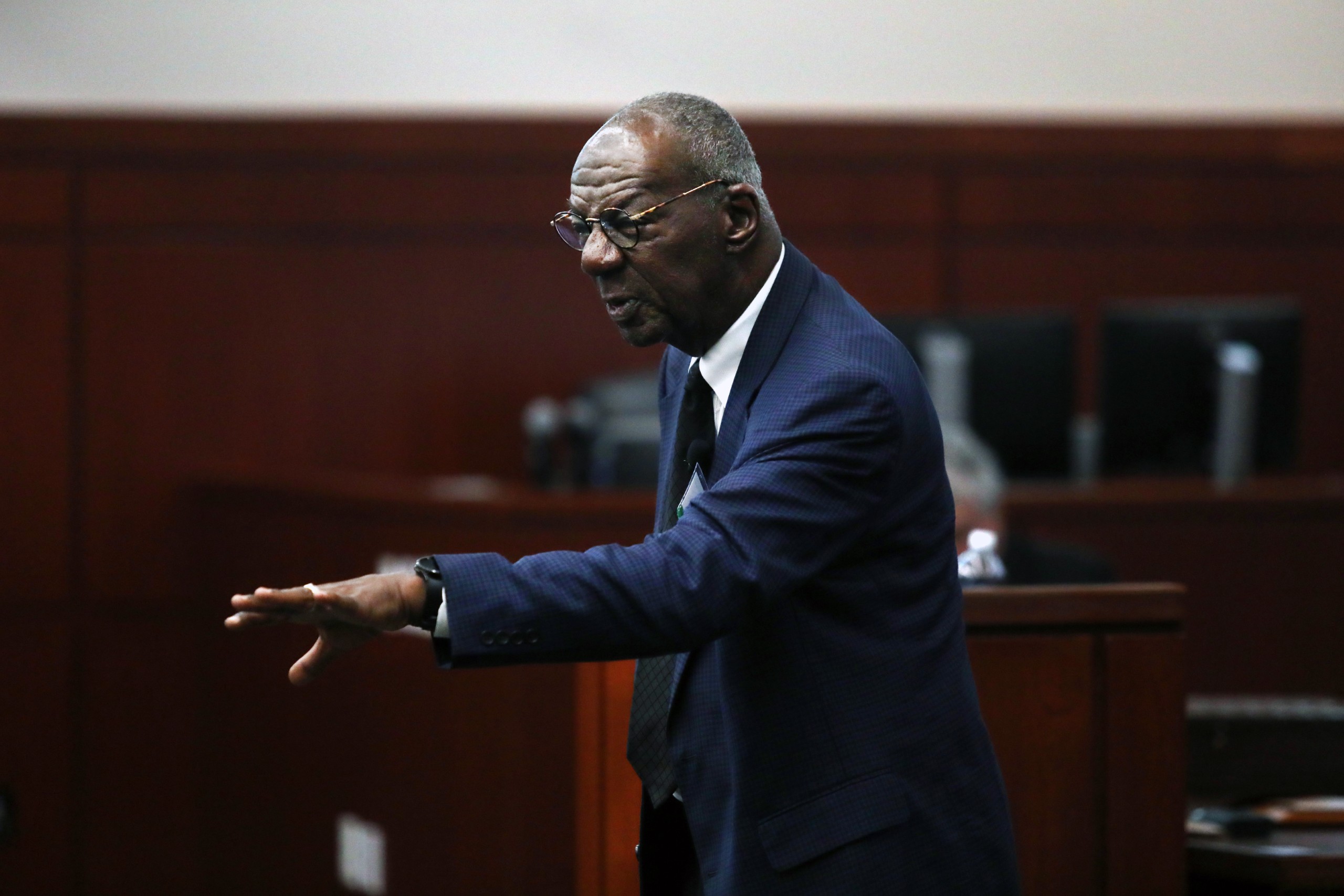 A person stands behind a lectern, gesturing while speaking to an audience in a courtroom setting.