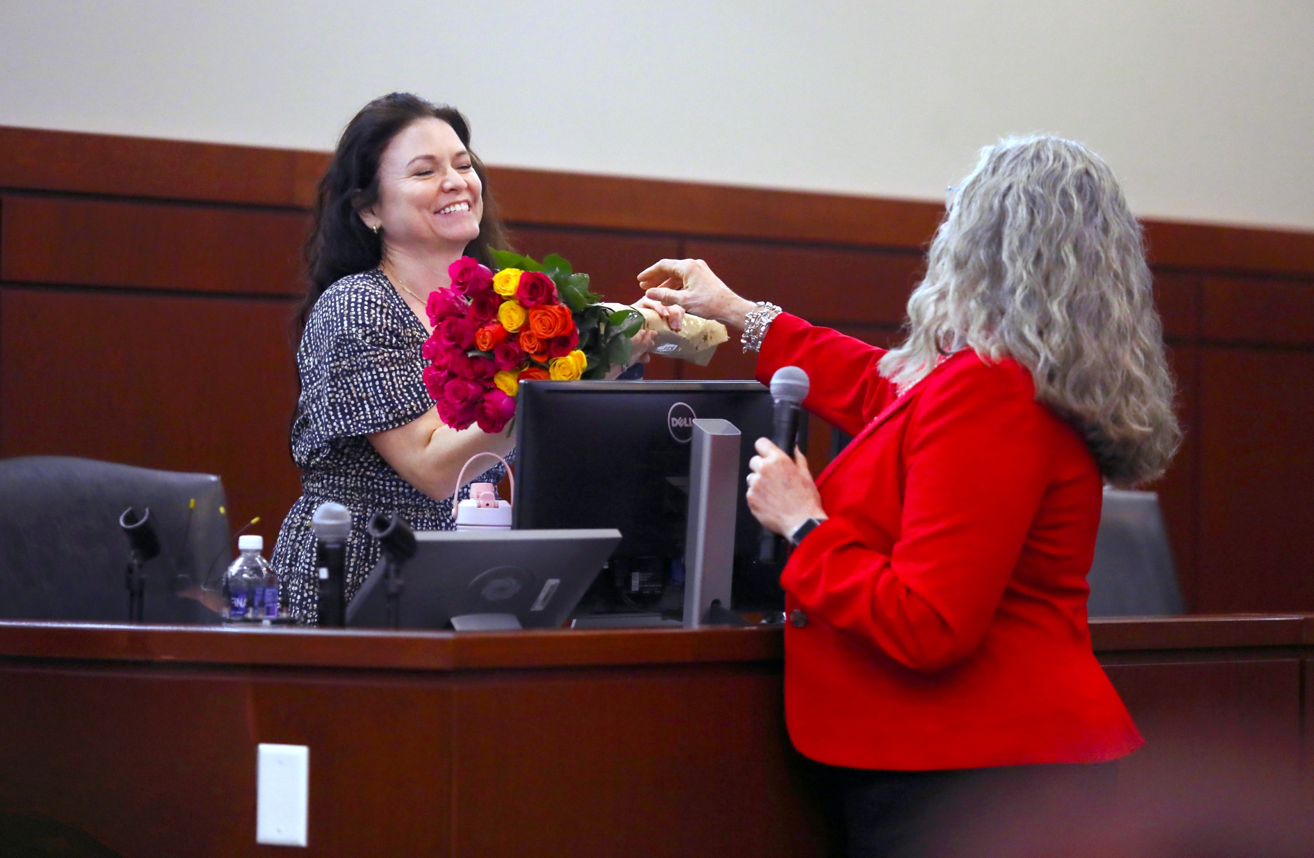Two individuals are interacting at a lectern in a formal setting. One person is handing a bouquet of flowers to the other, who is smiling in appreciation. There is a microphone and a computer on the lectern, and seating is visible in the background.