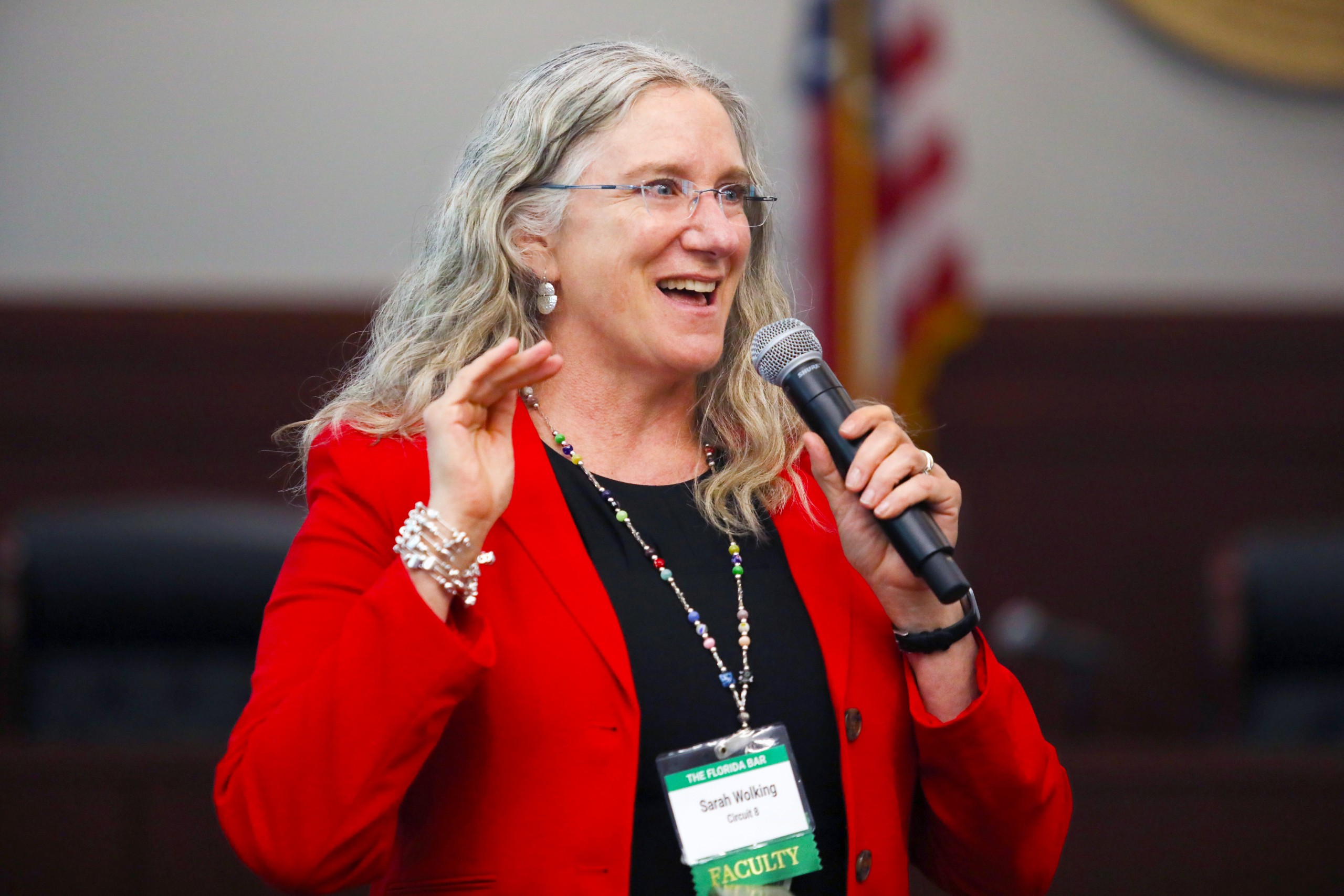 A person stands behind a lectern, speaking to an audience, smiling and gesturing with one hand while holding a microphone in the other. A name tag is visible on their outfit. In the background, chairs and an American flag can be seen.