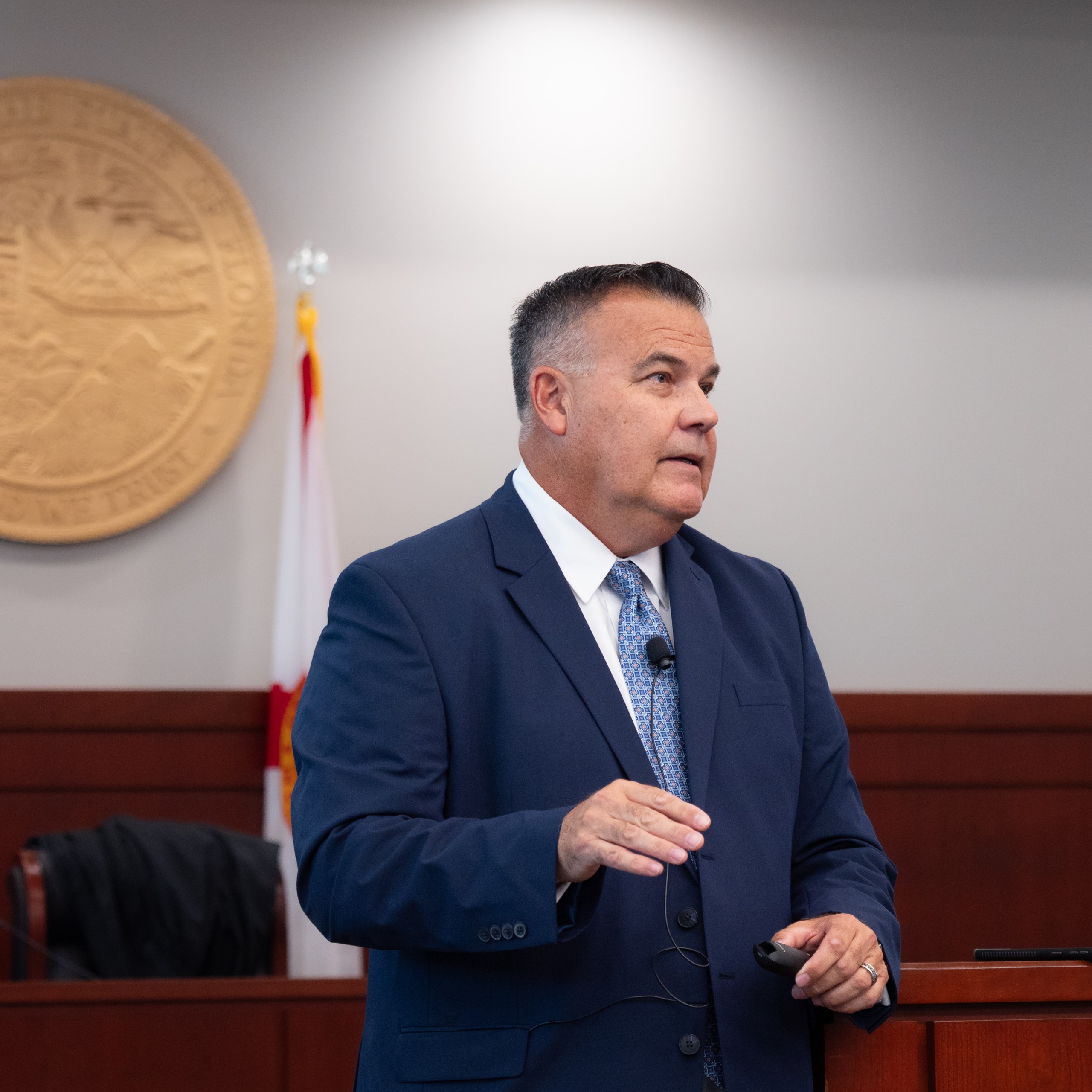 A person stands behind a lectern in a courtroom, speaking while gesturing. A state seal is visible on the wall behind them, and a chair is positioned in the background.