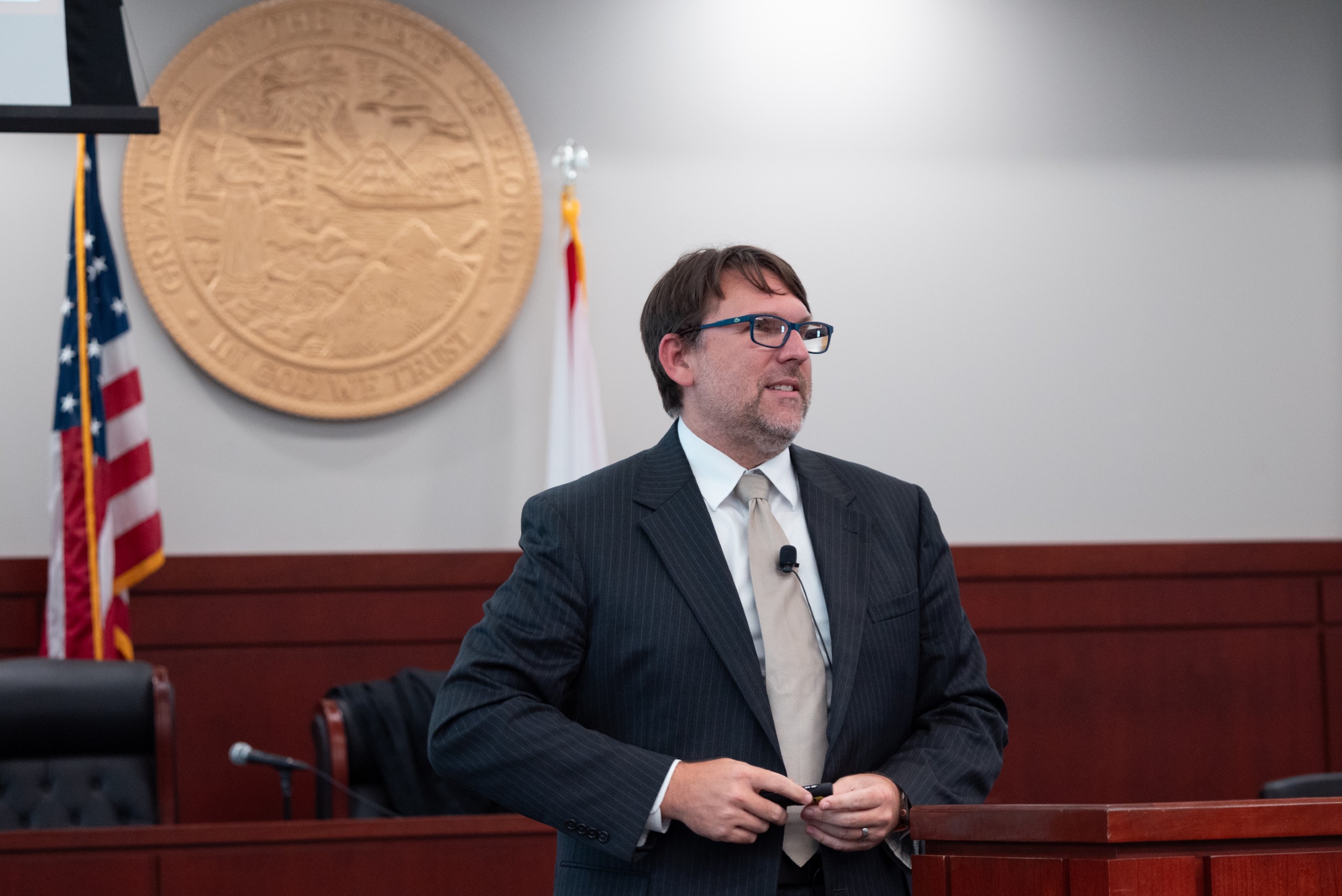 A person is speaking behind a lectern in a courtroom setting, with flags in the background and a state seal on the wall.