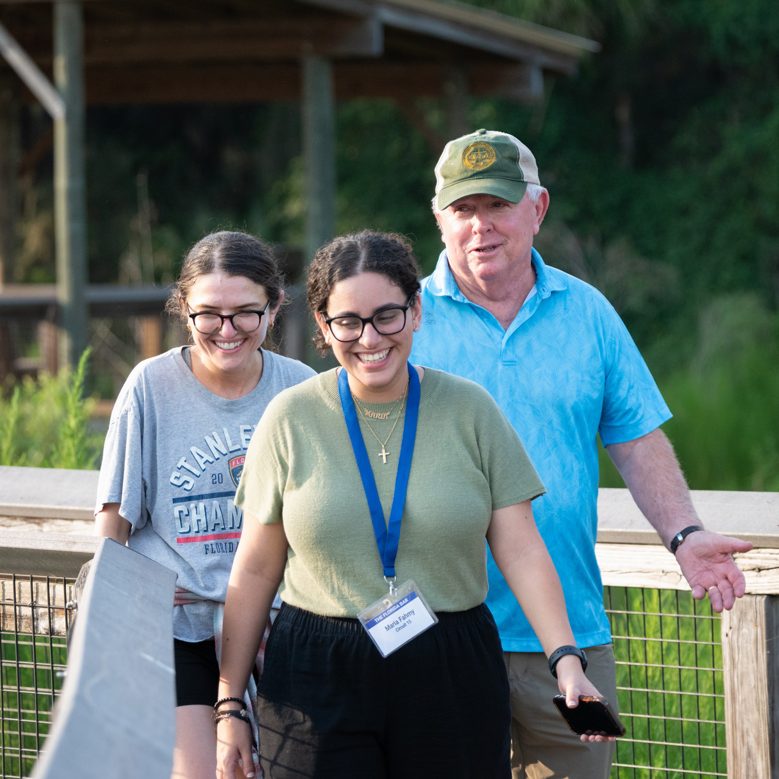 Three individuals are walking on a wooden pathway surrounded by greenery. Two are smiling while they walk, and one is gesturing with hands. The setting appears casual and outdoors.
