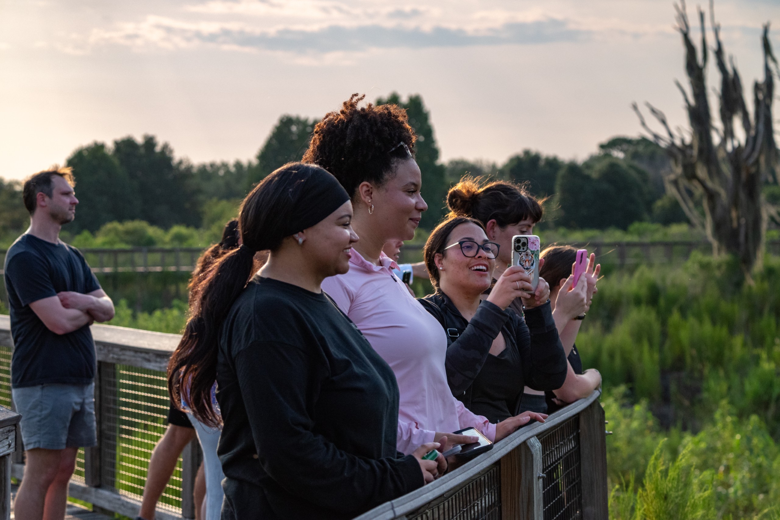 A group of people stands on a wooden boardwalk, enjoying the view of a natural landscape. Some individuals are smiling and taking photos, while others are looking out over the scenery. The setting appears to be during sunset, with greenery and trees in the background.