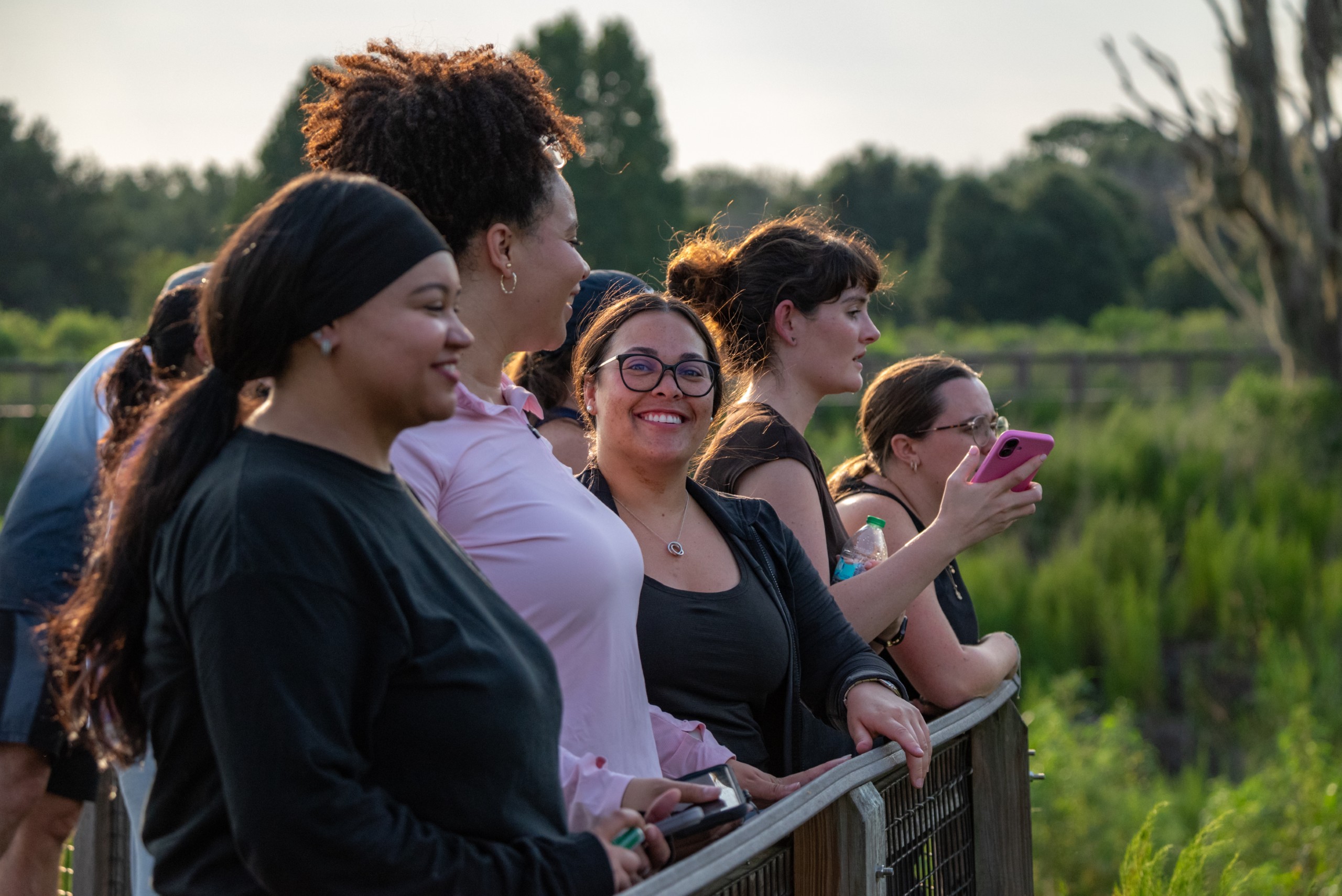 A group of individuals stands together along a railing, smiling and engaging with one another in a natural outdoor setting. Some are holding mobile phones, while others appear to be enjoying the moment. Lush greenery is visible in the background.