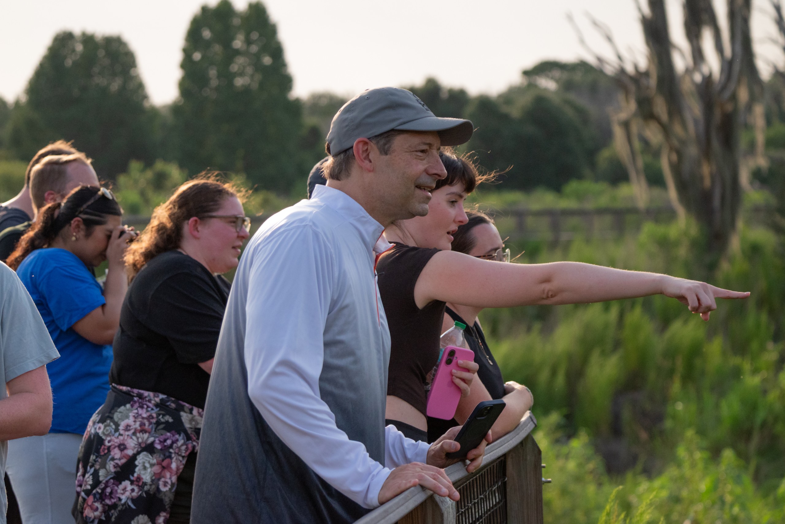 A group of people stands on a wooden railing, observing the scenery. One person points towards something in the distance while others appear engaged in conversation, enjoying the environment. Green foliage and trees are visible in the background.