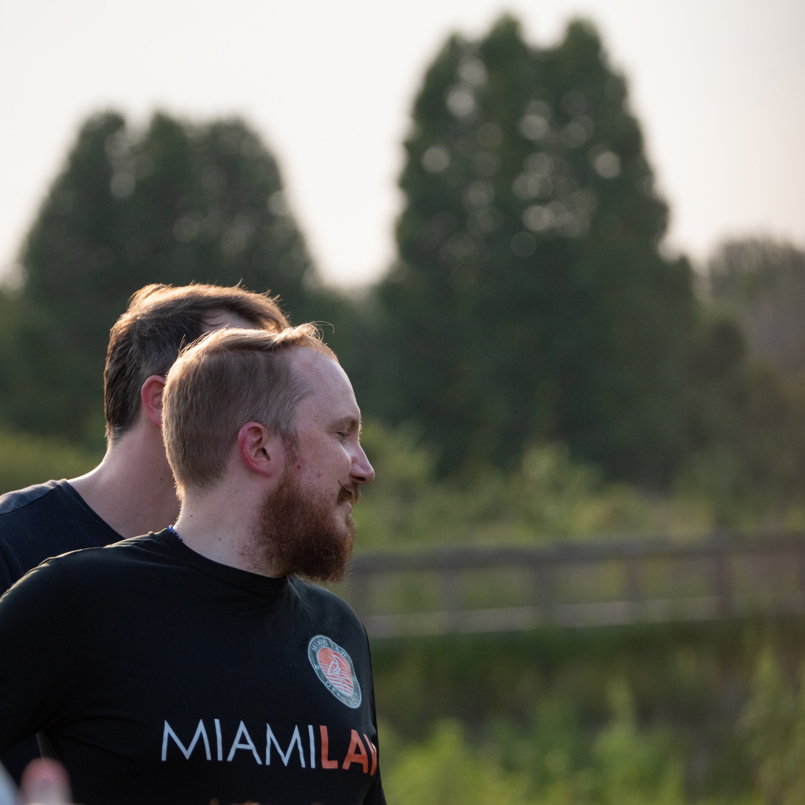 Two individuals are seen outdoors, with trees in the background. One person is smiling and facing to the side. The scene suggests a relaxed atmosphere, possibly during an outdoor gathering or event.
