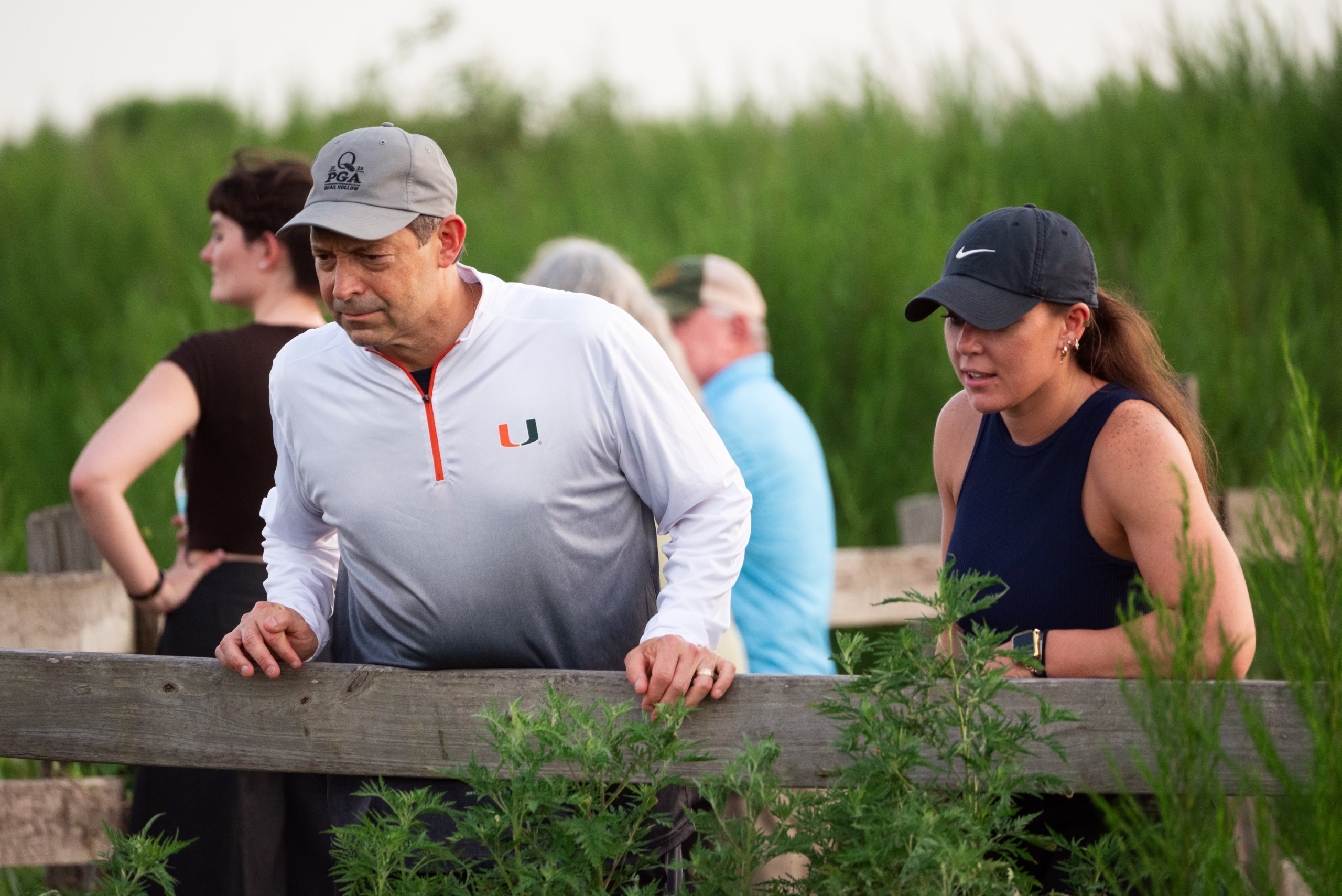 A group of individuals stands near a wooden fence, observing the landscape. Two people in the foreground lean on the fence, appearing engaged in conversation about their surroundings, while others are visible in the background. Lush greenery surrounds the scene.