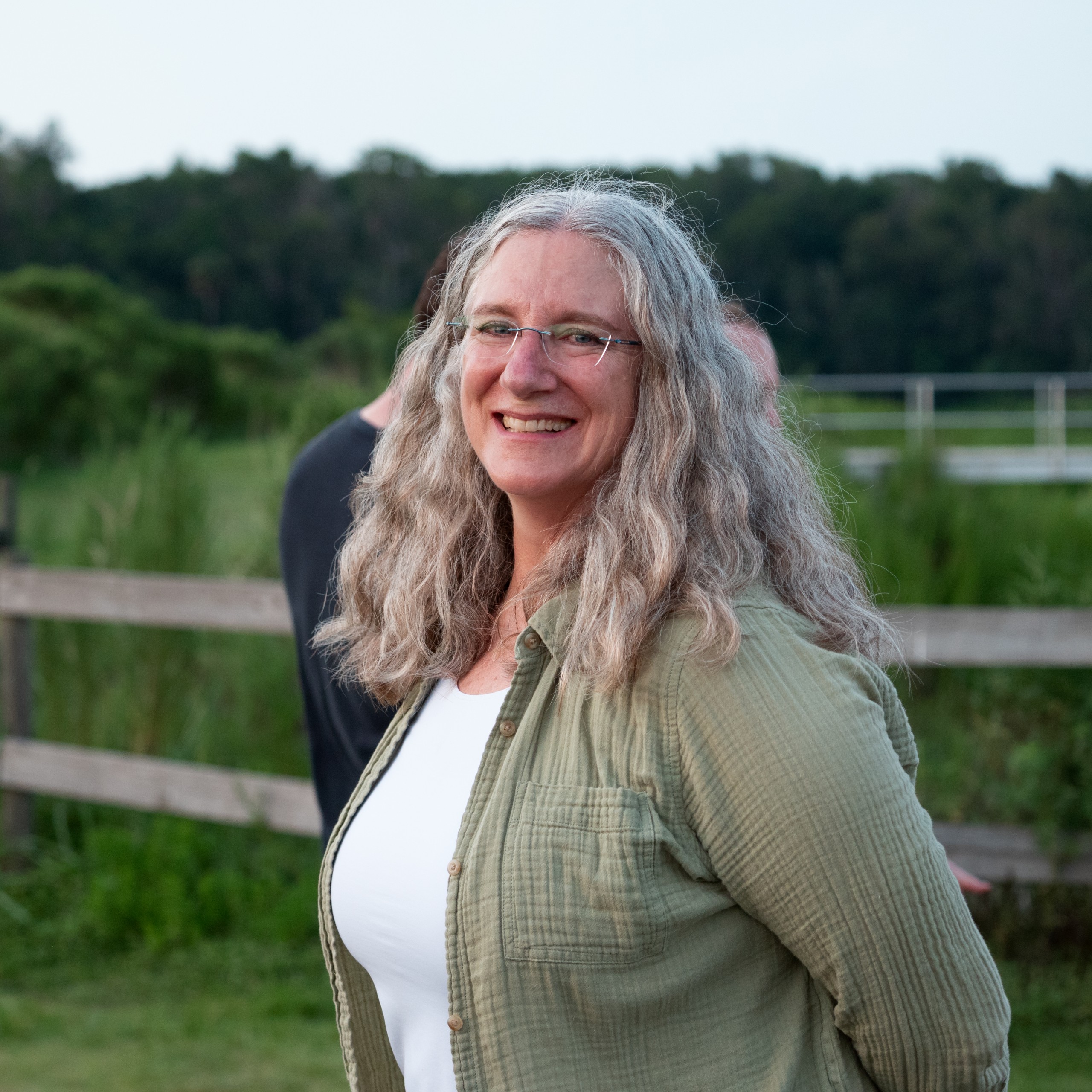 A person smiles while standing outdoors in front of a wooden fence, with green vegetation and trees in the background.