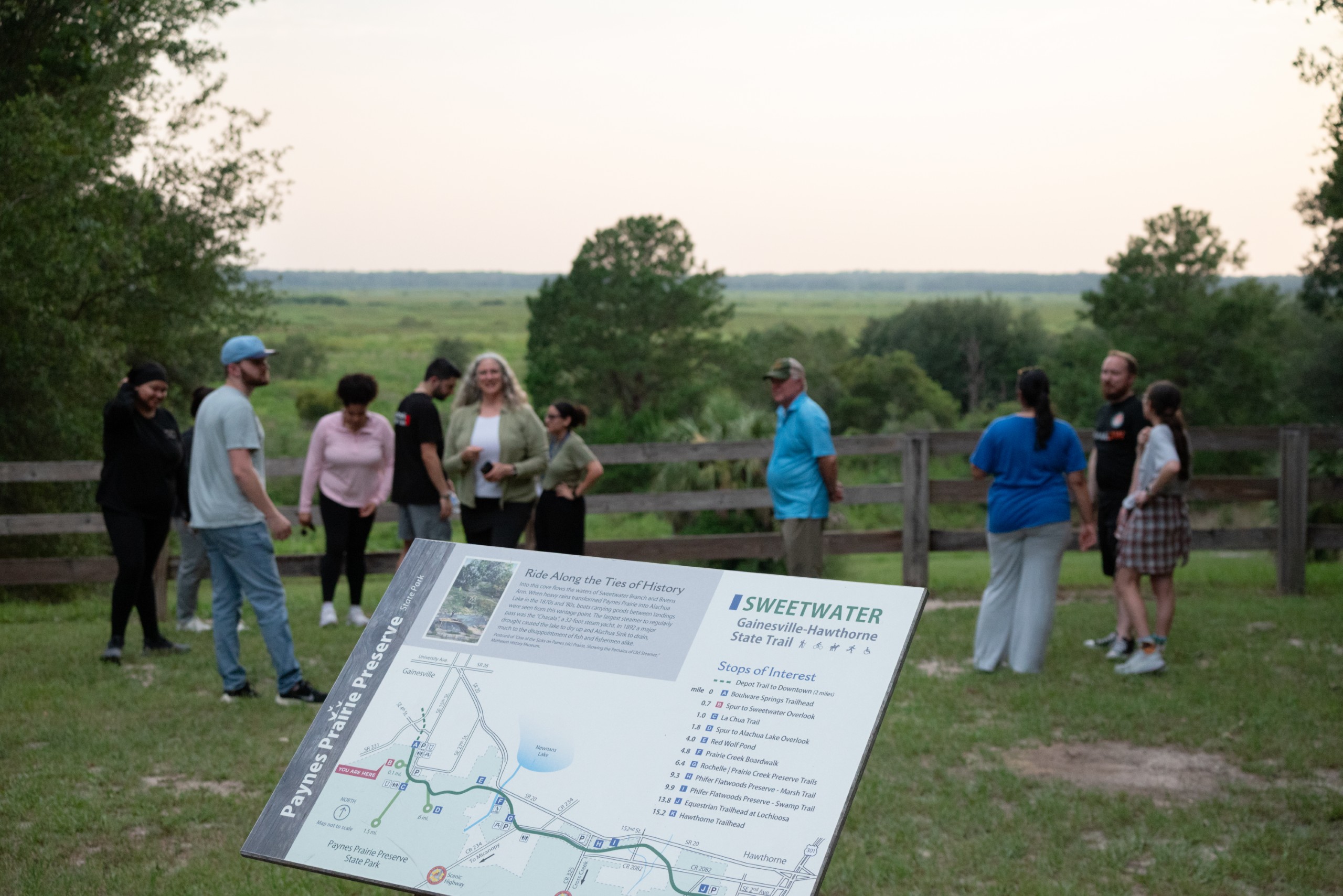 A group of people stands in a natural setting with trees and a large open field in the background. In the foreground, a map of the Paynes Prairie Preserve and the Sweetwater Gainesville-Hawthorne State Trail is prominently displayed, highlighting various points of interest.