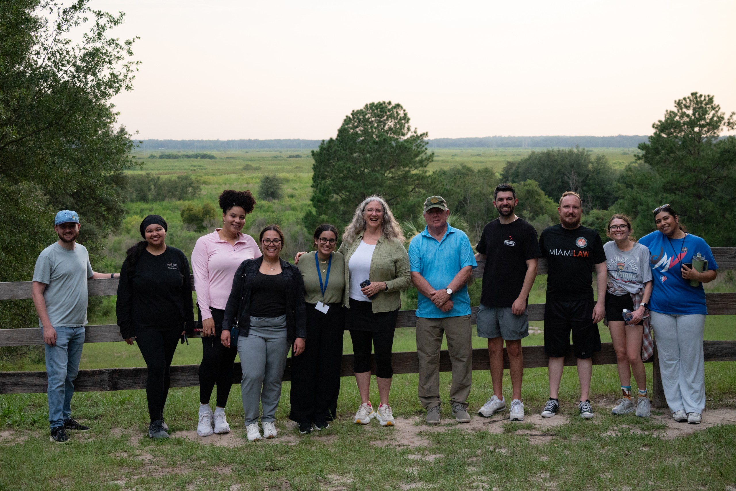 A group of ten people stands in a grassy area near a wooden fence, with a scenic view of trees and open fields in the background. They are arranged in two rows, smiling and posing for the photo.