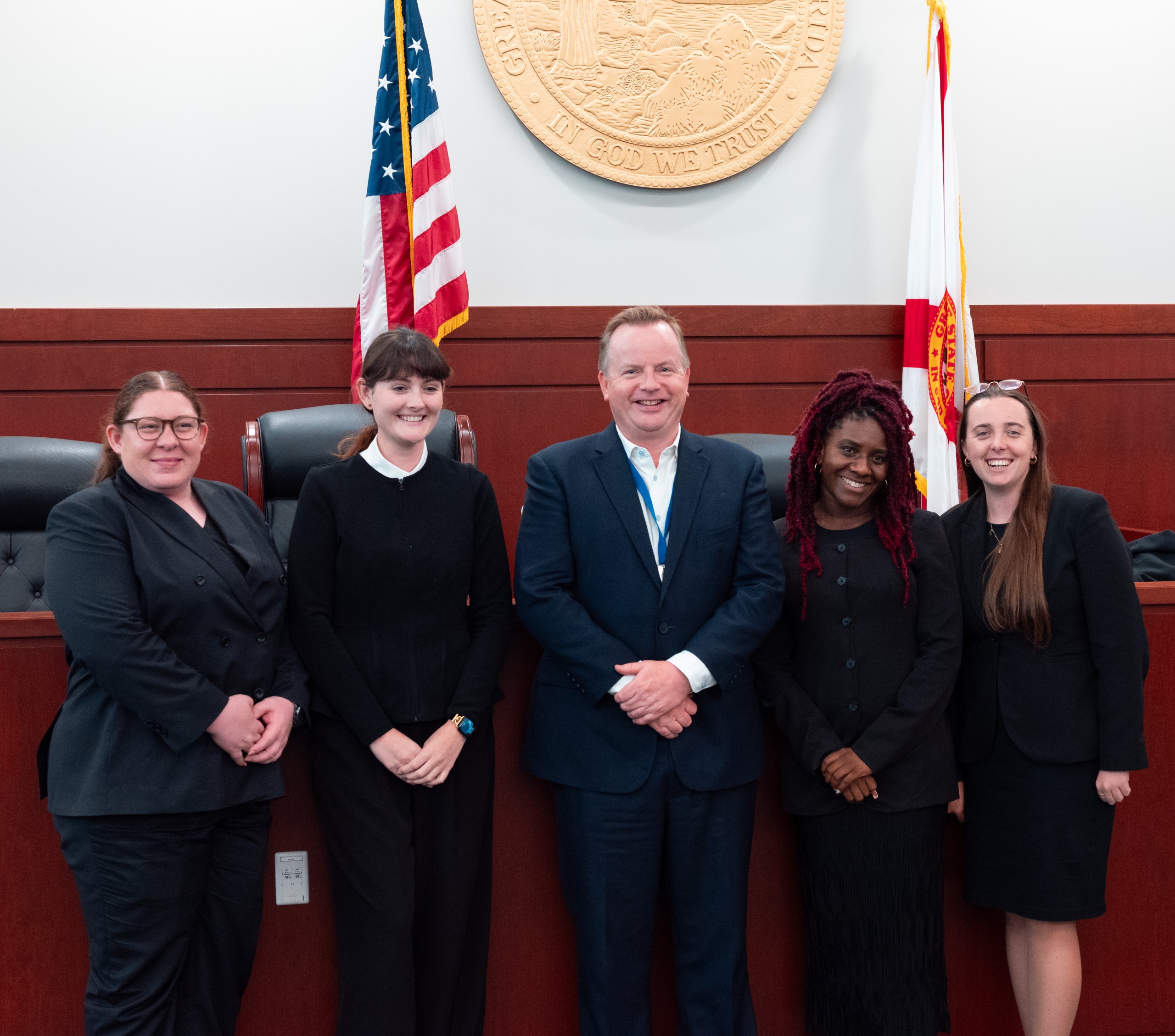 A group of five individuals stands together in a court setting, smiling. They are positioned in front of a wooden backdrop featuring a state seal and two flags. The group appears united and engaged, reflecting a professional atmosphere.