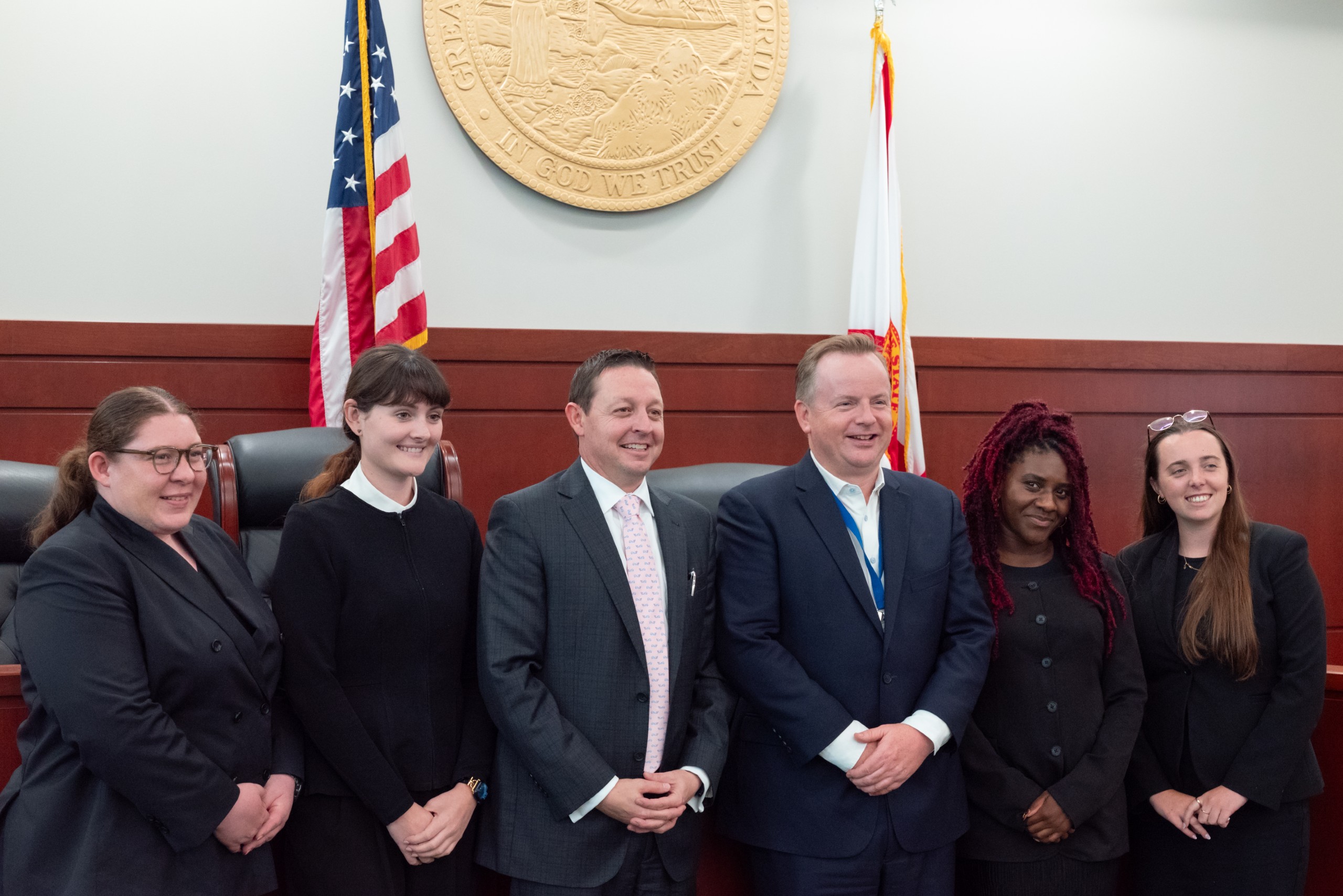 A group of six individuals stands together in a formal setting, smiling for the camera. An emblem representing Florida is displayed on the wall behind them, along with American flags. The group is arranged in two rows, with some individuals in front of others.