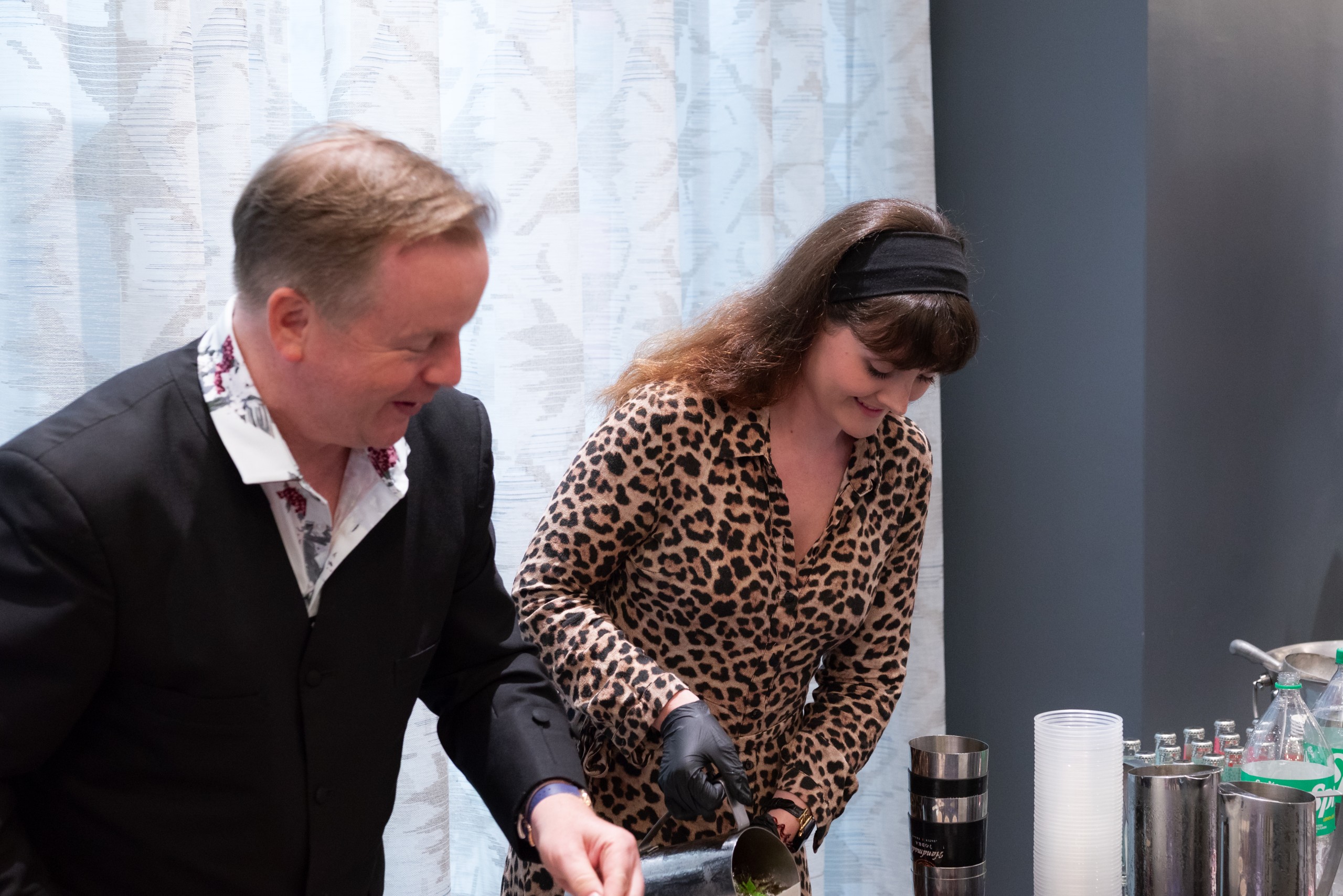 Two individuals are working together at a bar. One is pouring a drink, while the other is preparing ingredients. They are surrounded by various bar tools and containers. The background features a light patterned wall.