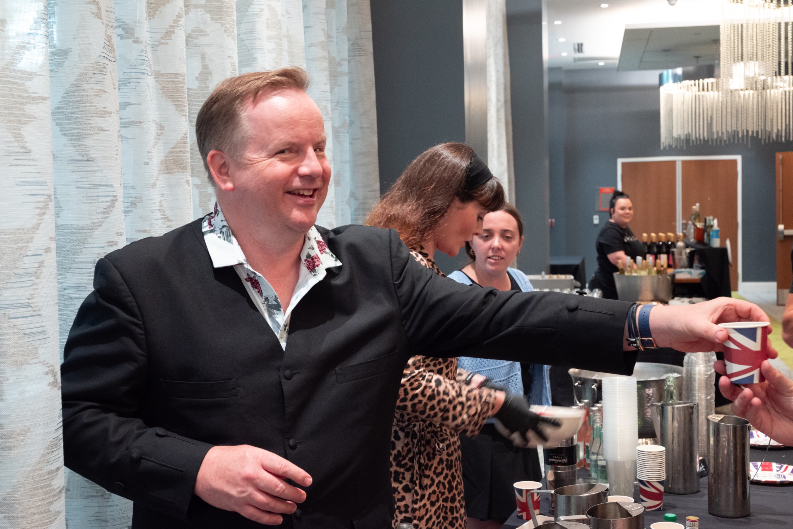 A group of individuals serves drinks at a bar. One person hands a cup to another while smiling, with various drink containers and a bar setup visible in the background.