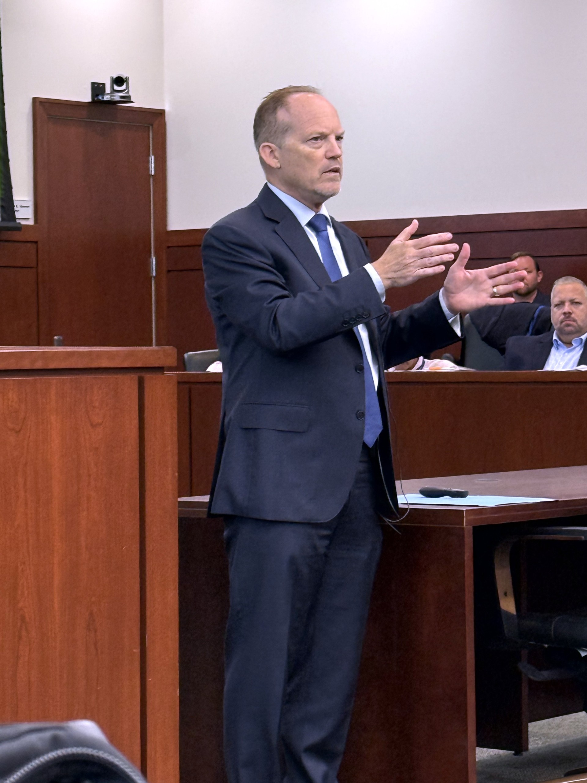 A person gestures while speaking in a courtroom setting, with an audience visible in the background.