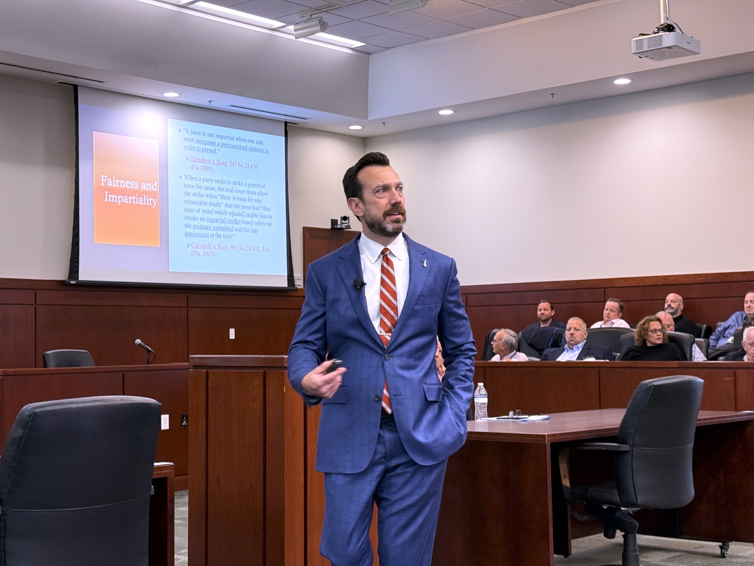 A person is standing behind a lectern while speaking to an audience in a courtroom setting. A presentation slide titled "Fairness and Impartiality" is visible on a screen behind them. Audience members are seated and listening attentively.