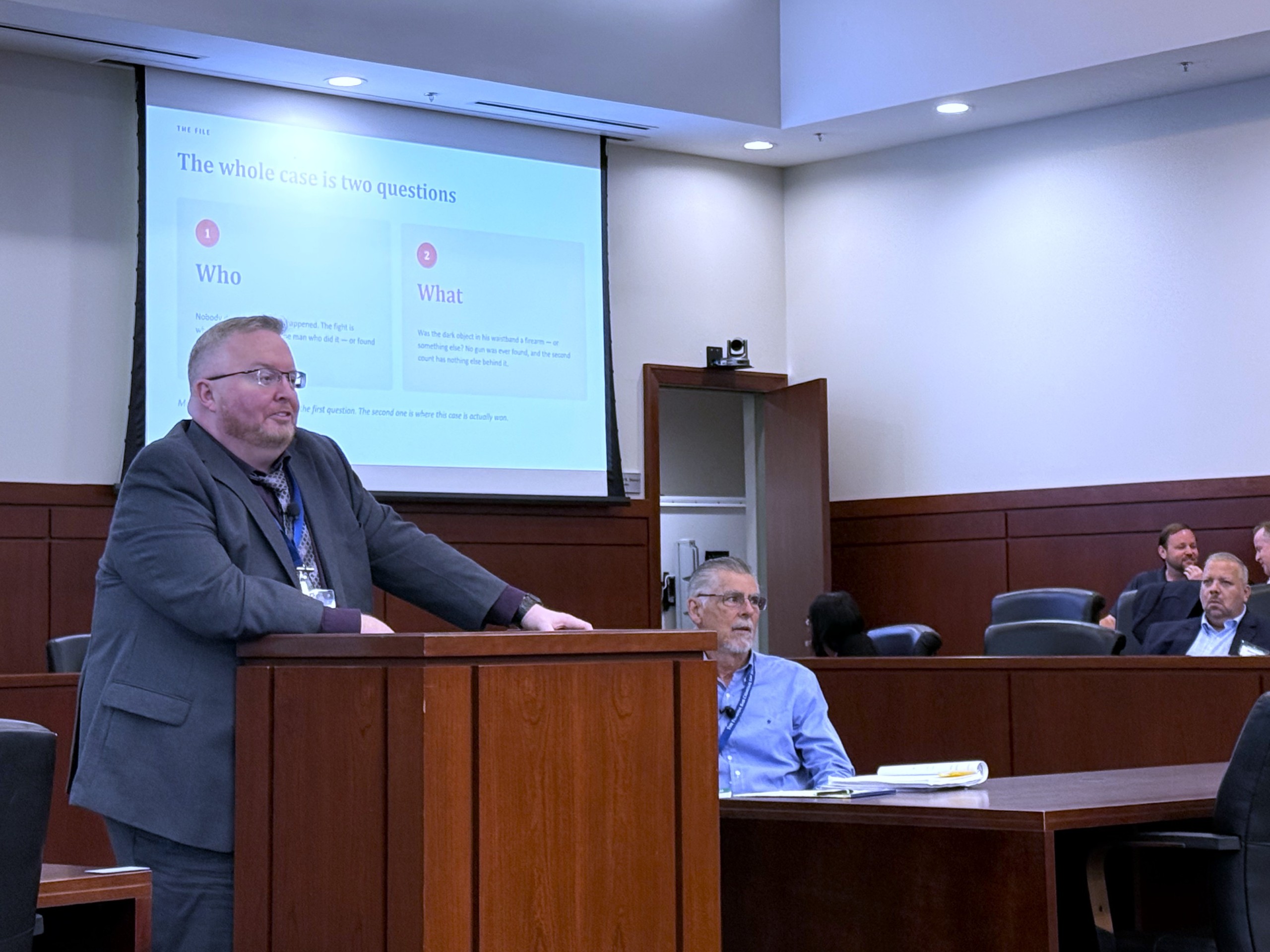 A person stands behind a lectern, speaking to an audience in a formal setting. A presentation slide is visible in the background with the text "The whole case is two questions: Who? What?" Several individuals are seated in the audience, engaged in the presentation.