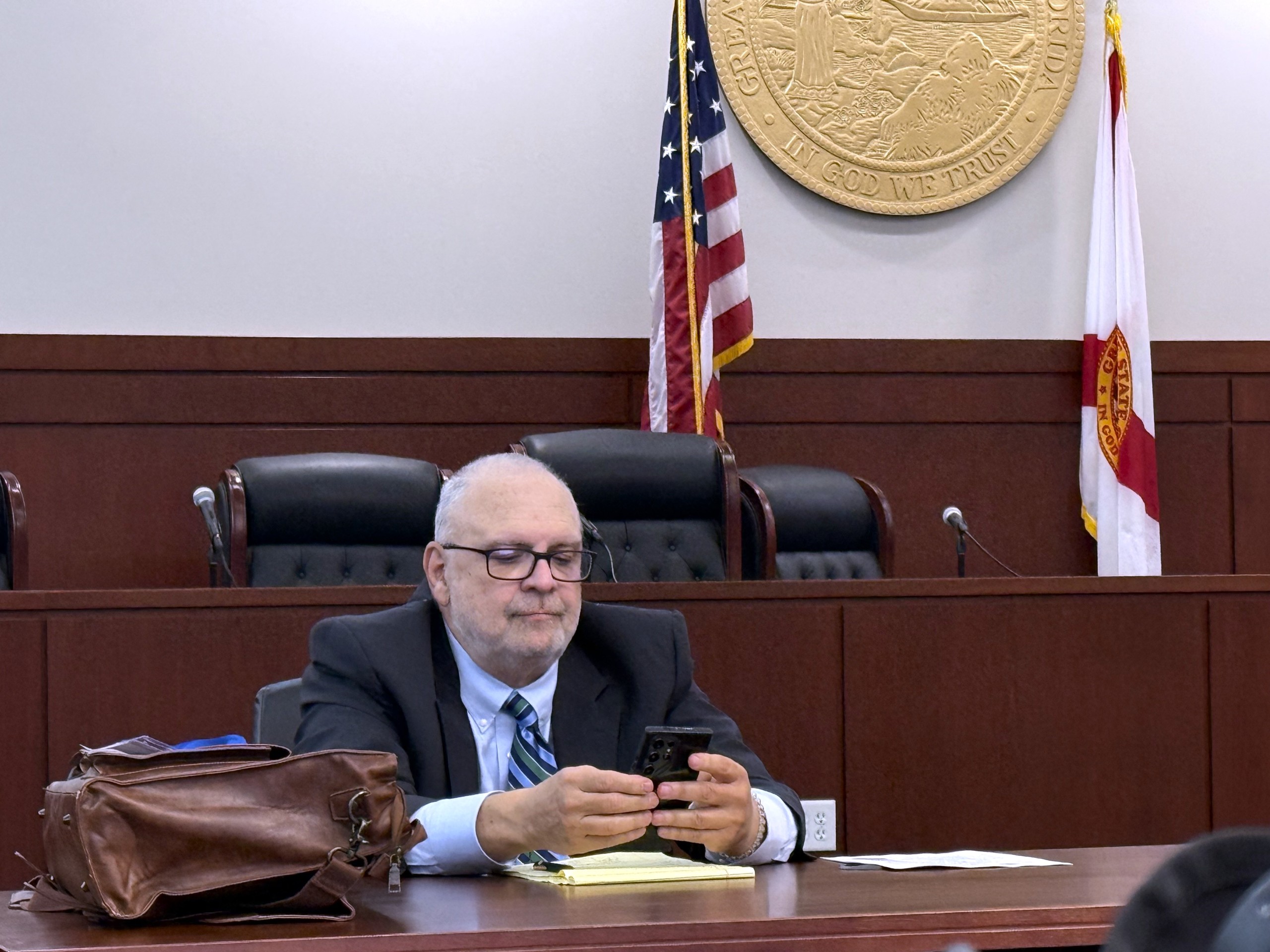 A person sits at a table in a courtroom, looking at a phone. There are empty chairs in the background, and flags and a state seal are visible on the wall behind. Items on the table include a notebook and a bag.