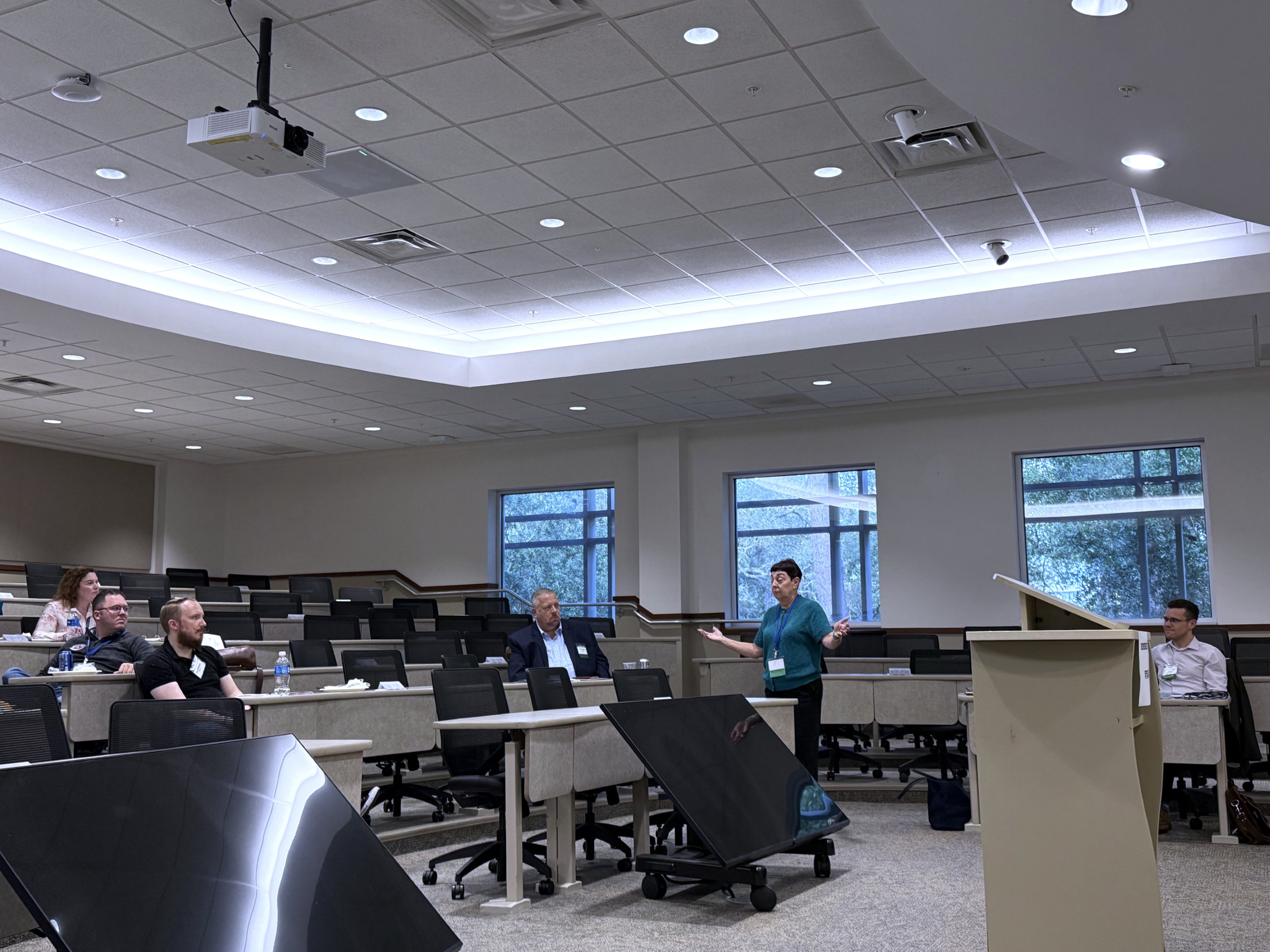 An individual is standing behind a lectern and speaking to an audience in a large, well-lit room. Several people are seated at tables, paying attention to the speaker. Windows in the background show greenery outside.