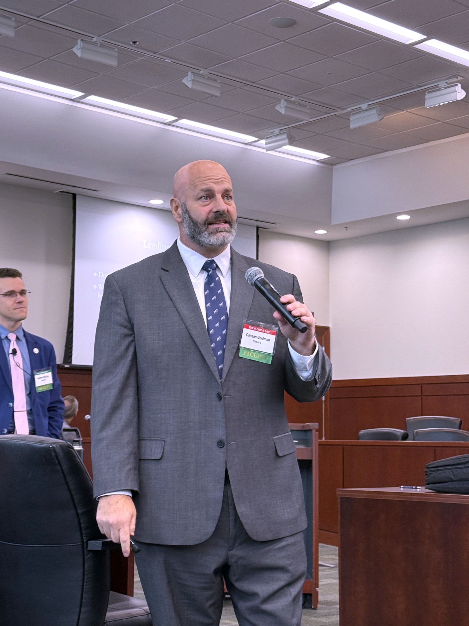 An individual stands behind a lectern, holding a microphone and speaking to an audience. In the background, a presentation screen is visible, and several seated attendees can be seen.