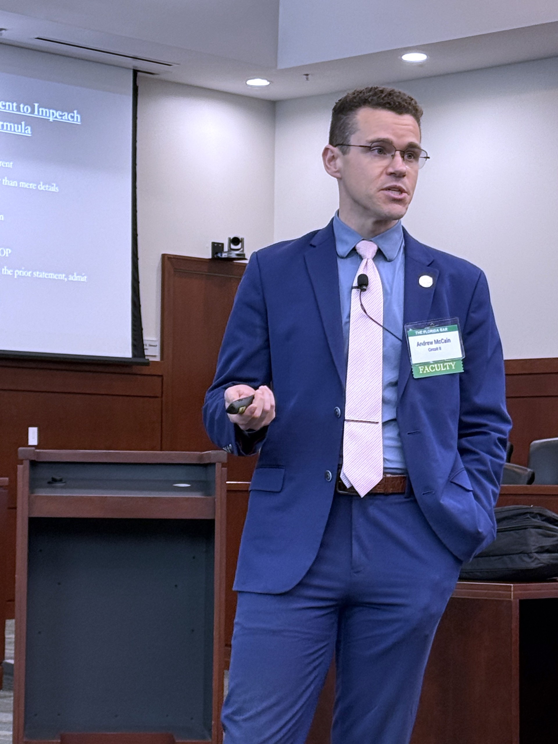 A person is speaking behind a lectern in a conference room, with a presentation displayed on a screen in the background. The speaker holds a microphone and gestures while presenting information to an audience.