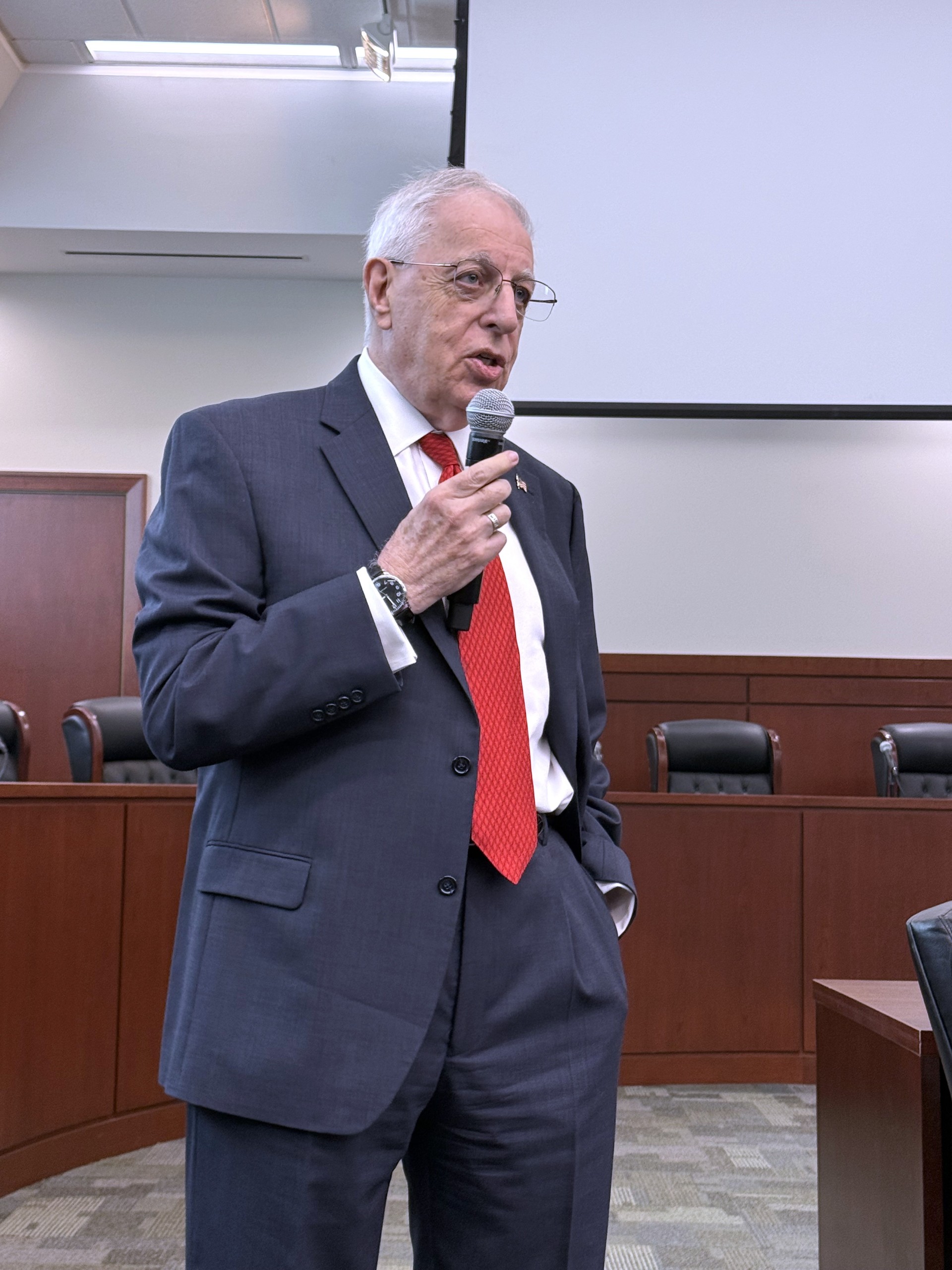 An individual speaks while holding a microphone, addressing an audience in a formal setting. A blank projector screen is visible in the background, and chairs are arranged for the attendees.