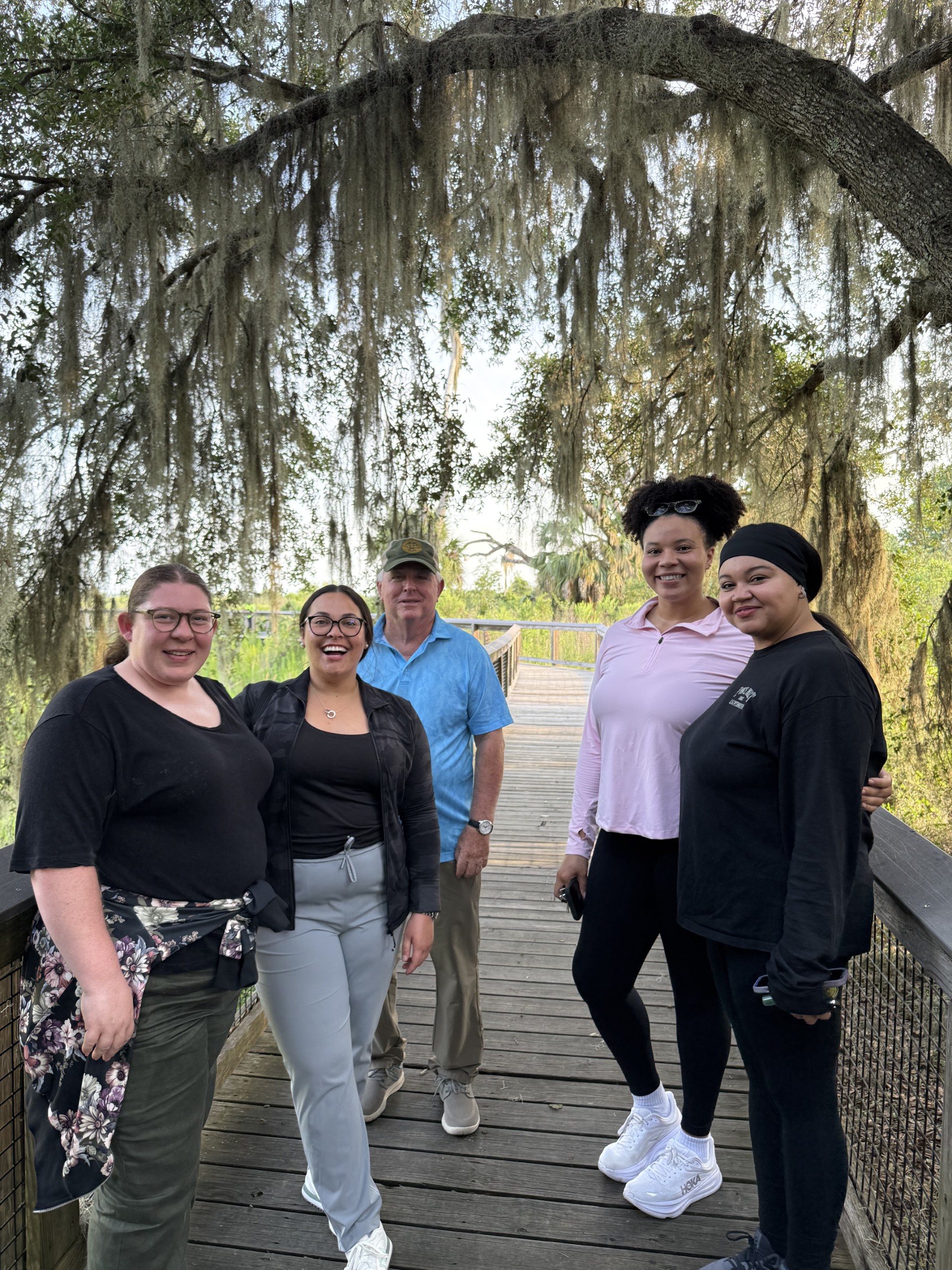 A group of five individuals stands together on a wooden walkway surrounded by trees. The scene features lush greenery and Spanish moss hanging from the branches above.