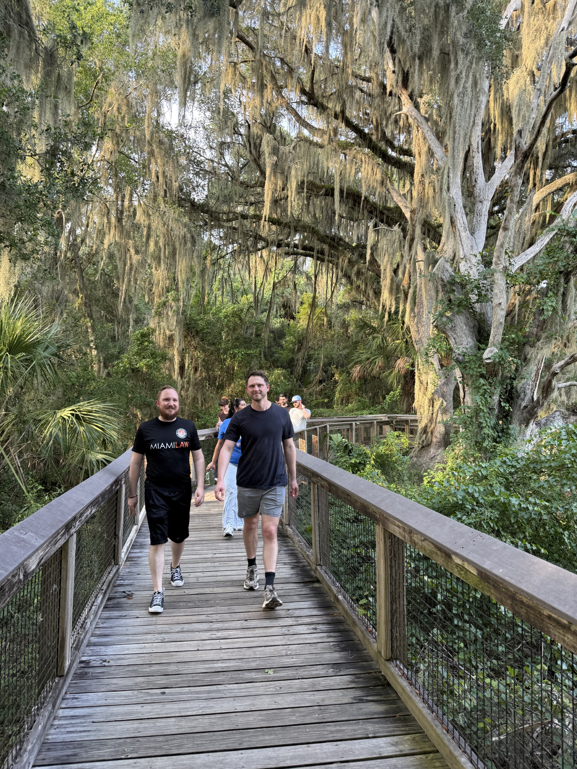 Two individuals walk on a wooden path through a lush, green area with trees and hanging moss. Additional people are walking in the background.