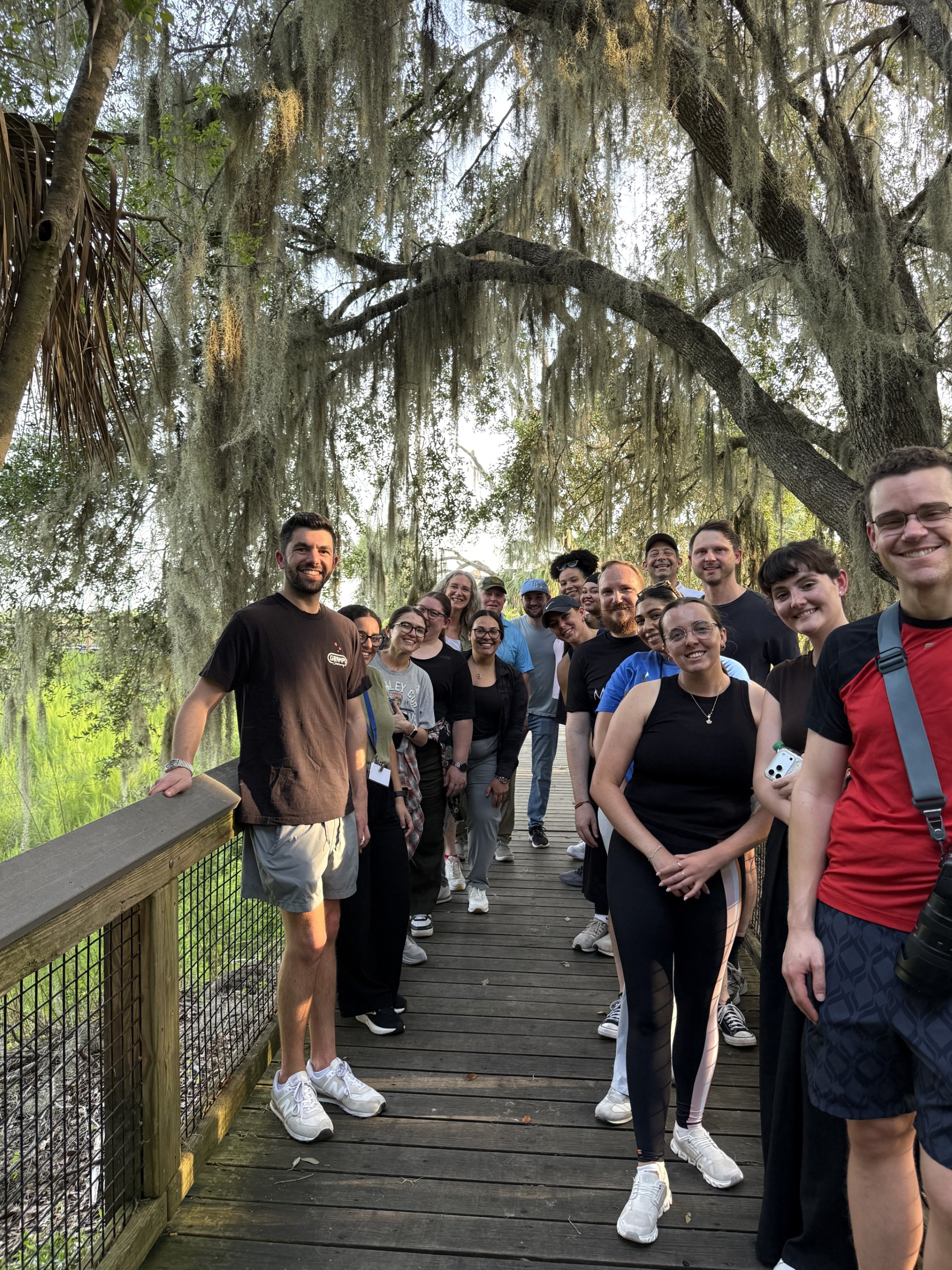 A group of individuals stands on a wooden boardwalk surrounded by trees. They are smiling and posing for the camera, with some people in the front and others in the background. Sunlight filters through the leaves above.