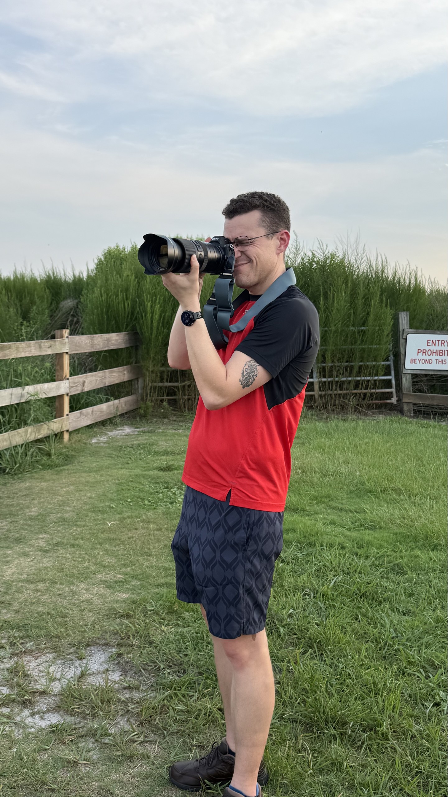 A person stands in a grassy area holding a camera with a large lens, preparing to take a photograph. Behind them, there is a wooden fence and tall grass in the background under a lightly clouded sky.