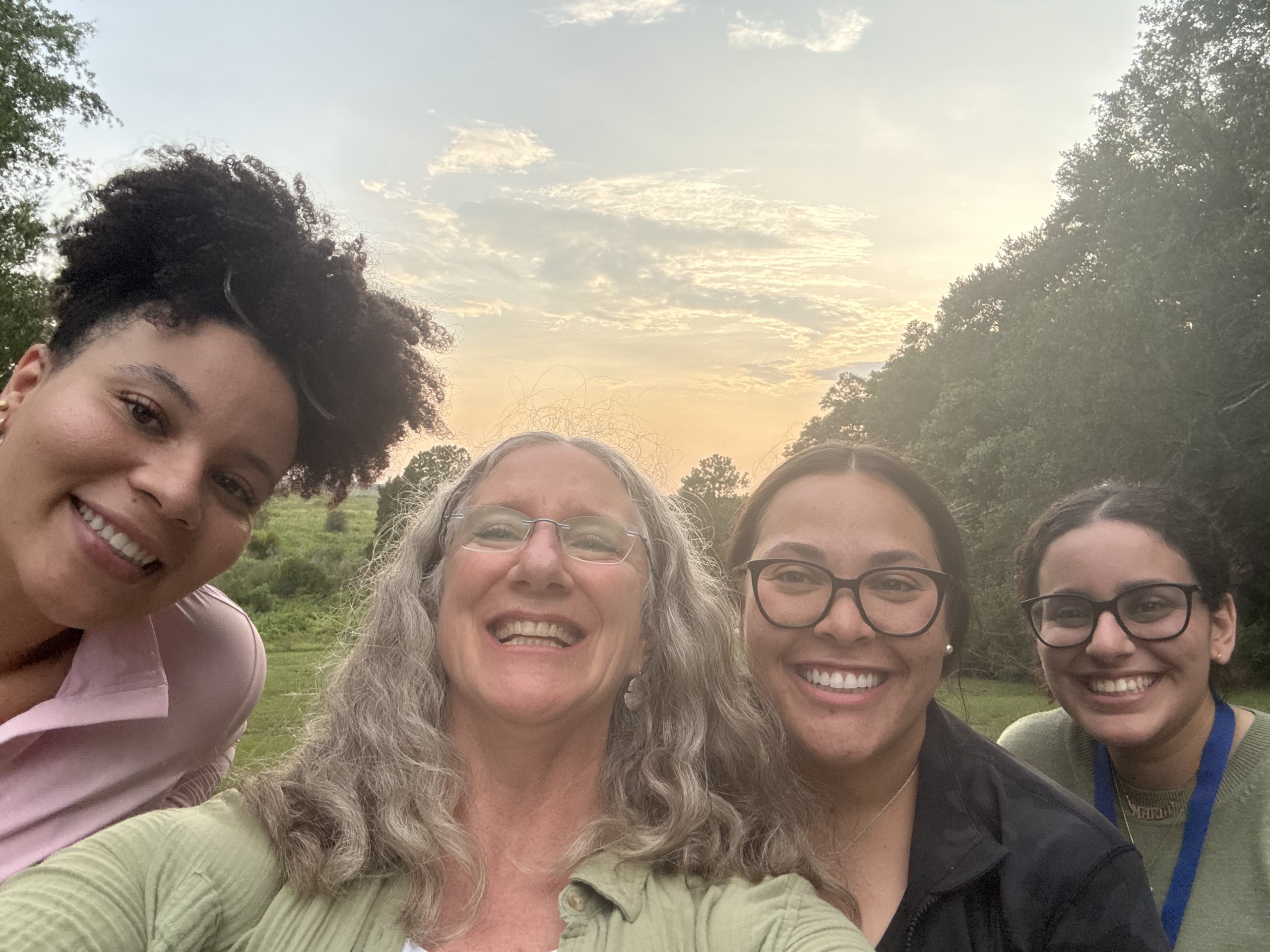 A group of four individuals poses for a selfie outdoors, with a sunset visible in the background. They are smiling and appear to be enjoying their time together in a natural setting.