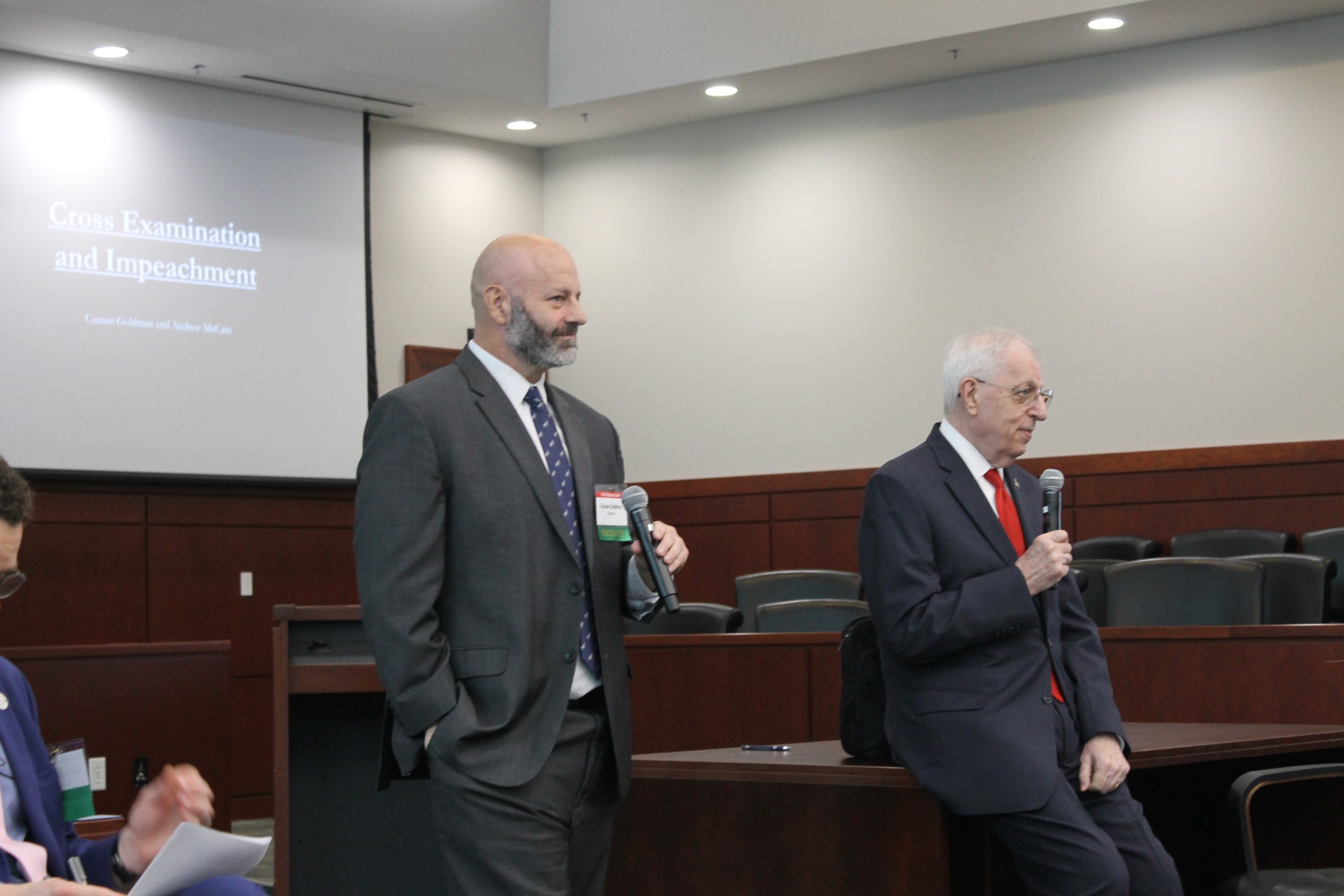 Two individuals are speaking in a courtroom setting. One is standing behind a lectern with a microphone, while the other is seated, also holding a microphone. A presentation on "Cross Examination and Impeachment" is visible on a screen in the background.