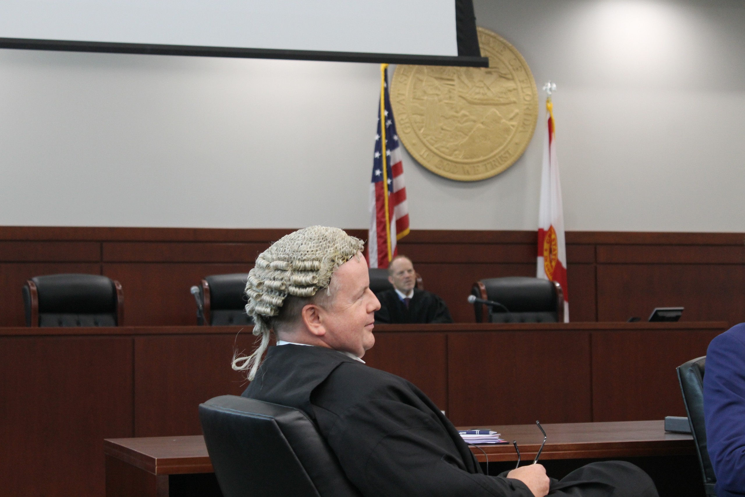A person wearing a wig sits in a courtroom, facing the audience. In the background, a judge is seated at a bench, with flags and a state seal visible on the wall.